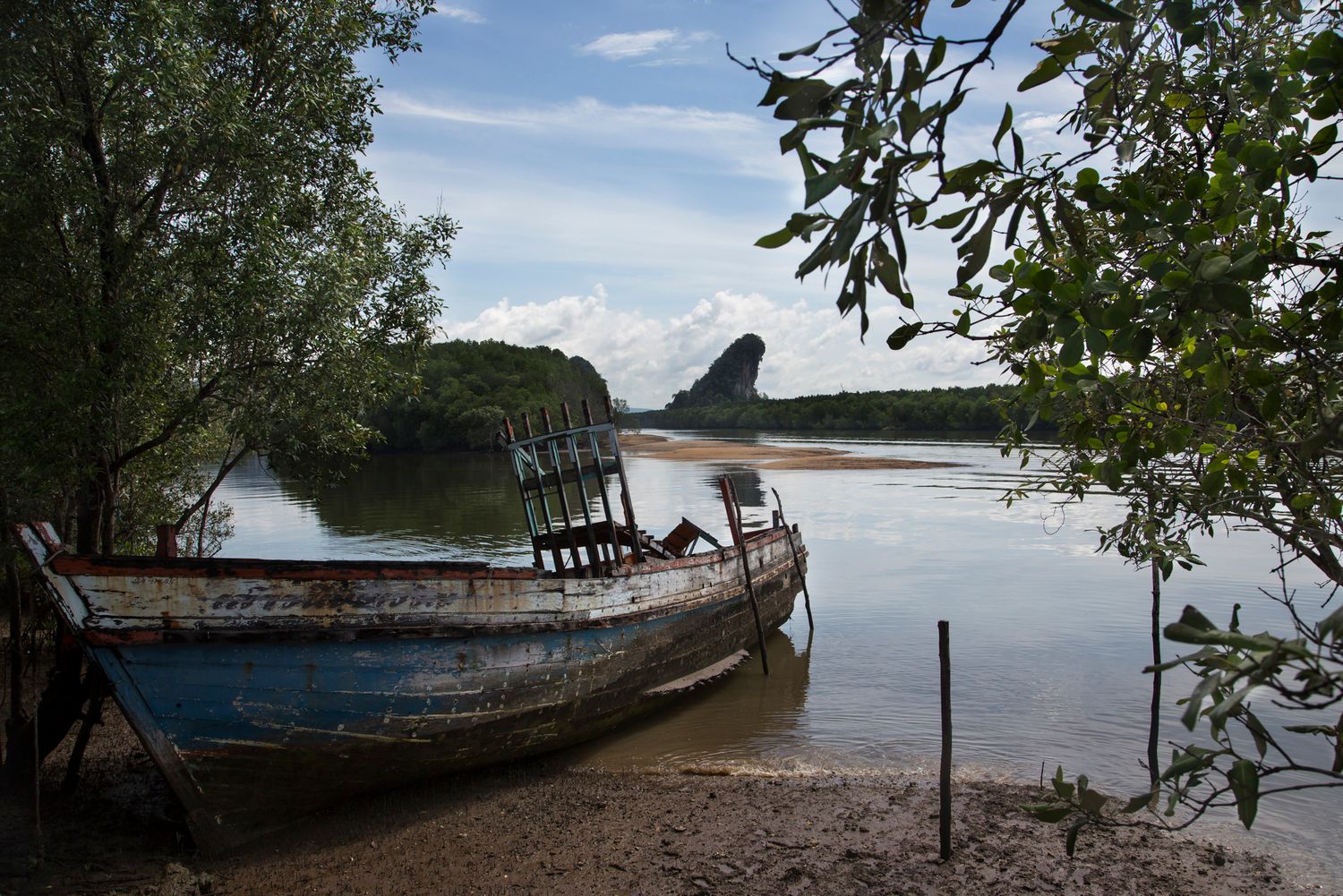 Old abandoned fishing boat resting on muddy riverbank surrounded by trees.