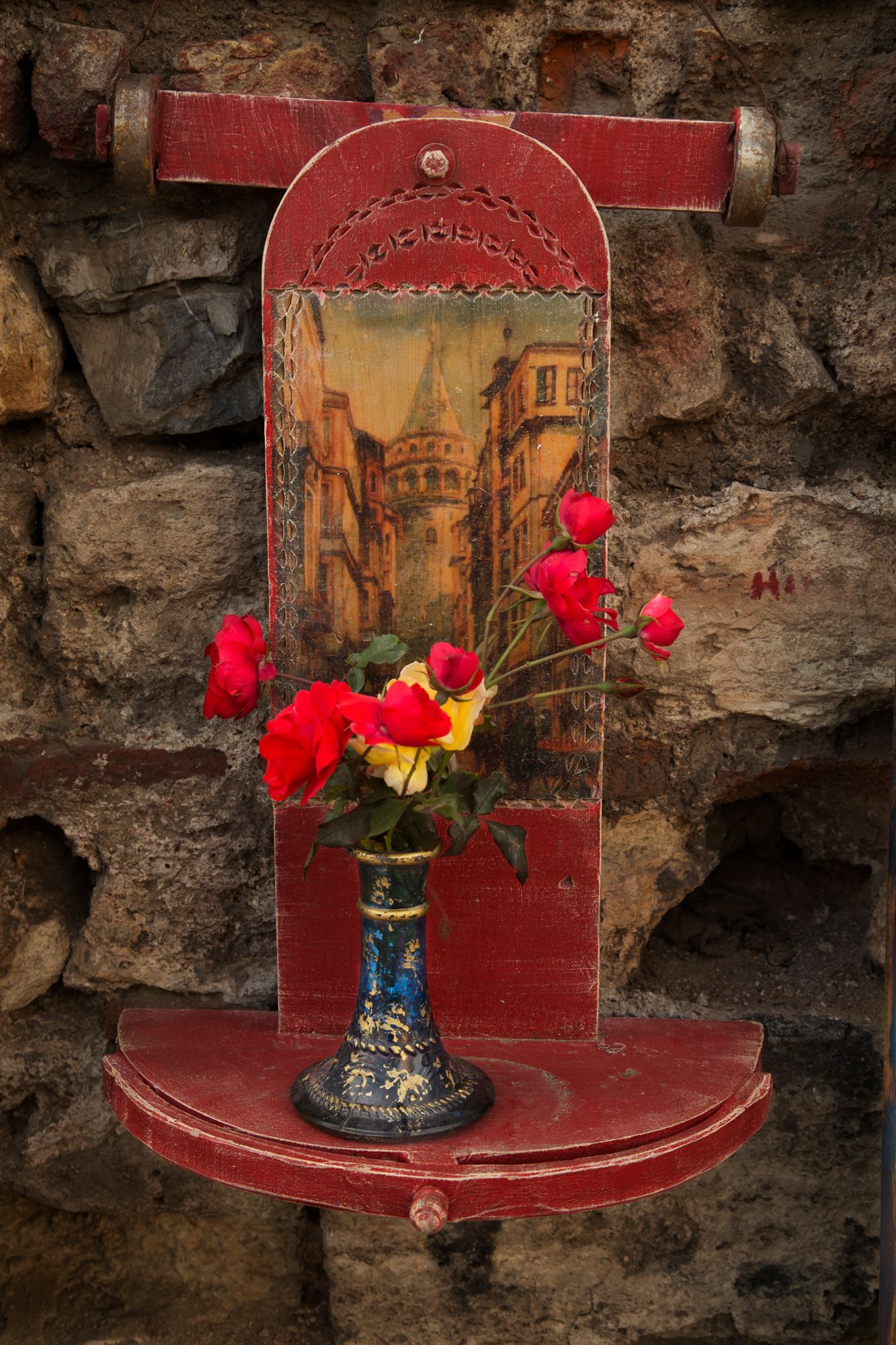 Vintage red wooden shelf with Italian street scene artwork and bright red flowers in blue vase against stone wall.