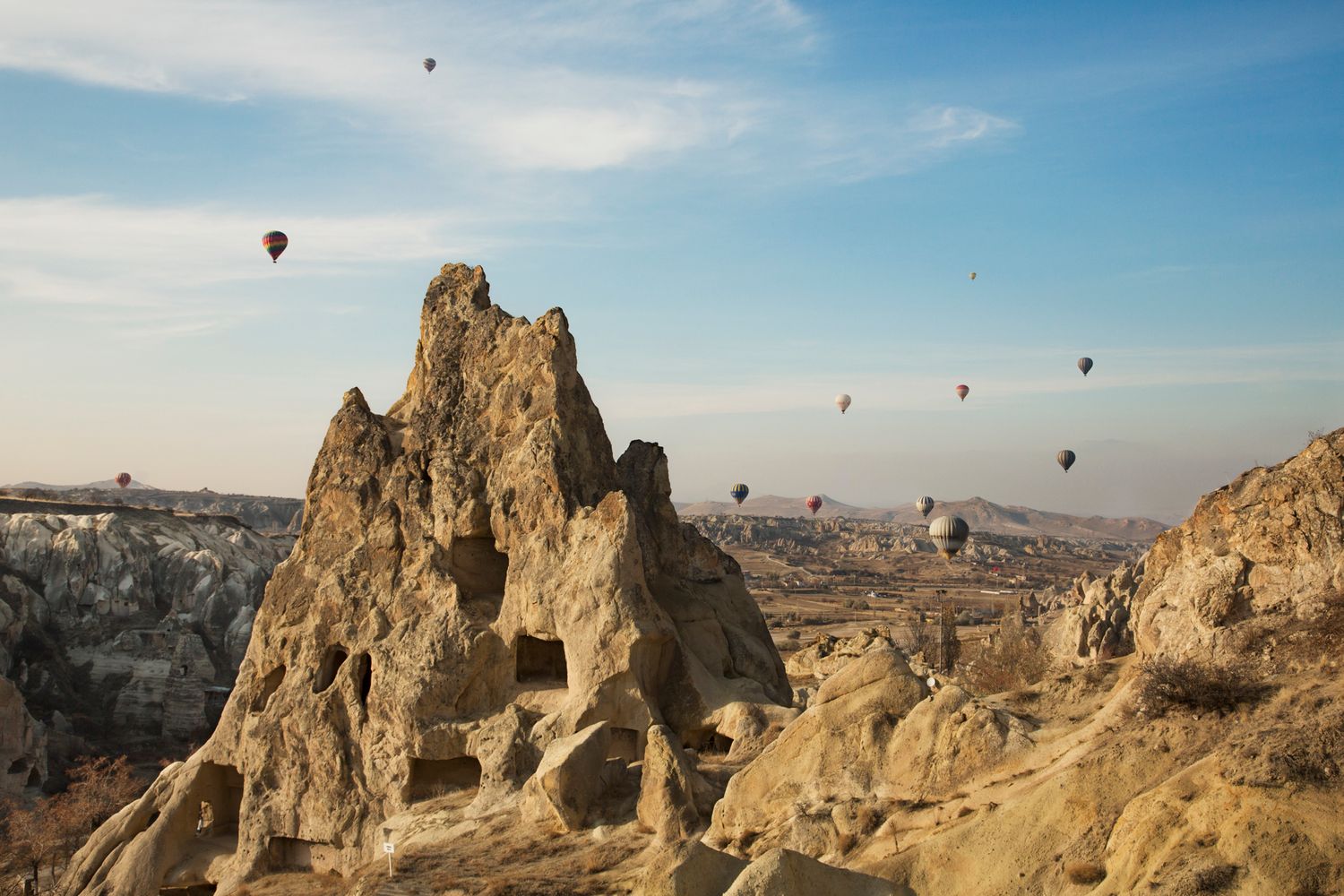 Hot air balloons float above rocky desert terrain against a bright blue sky.