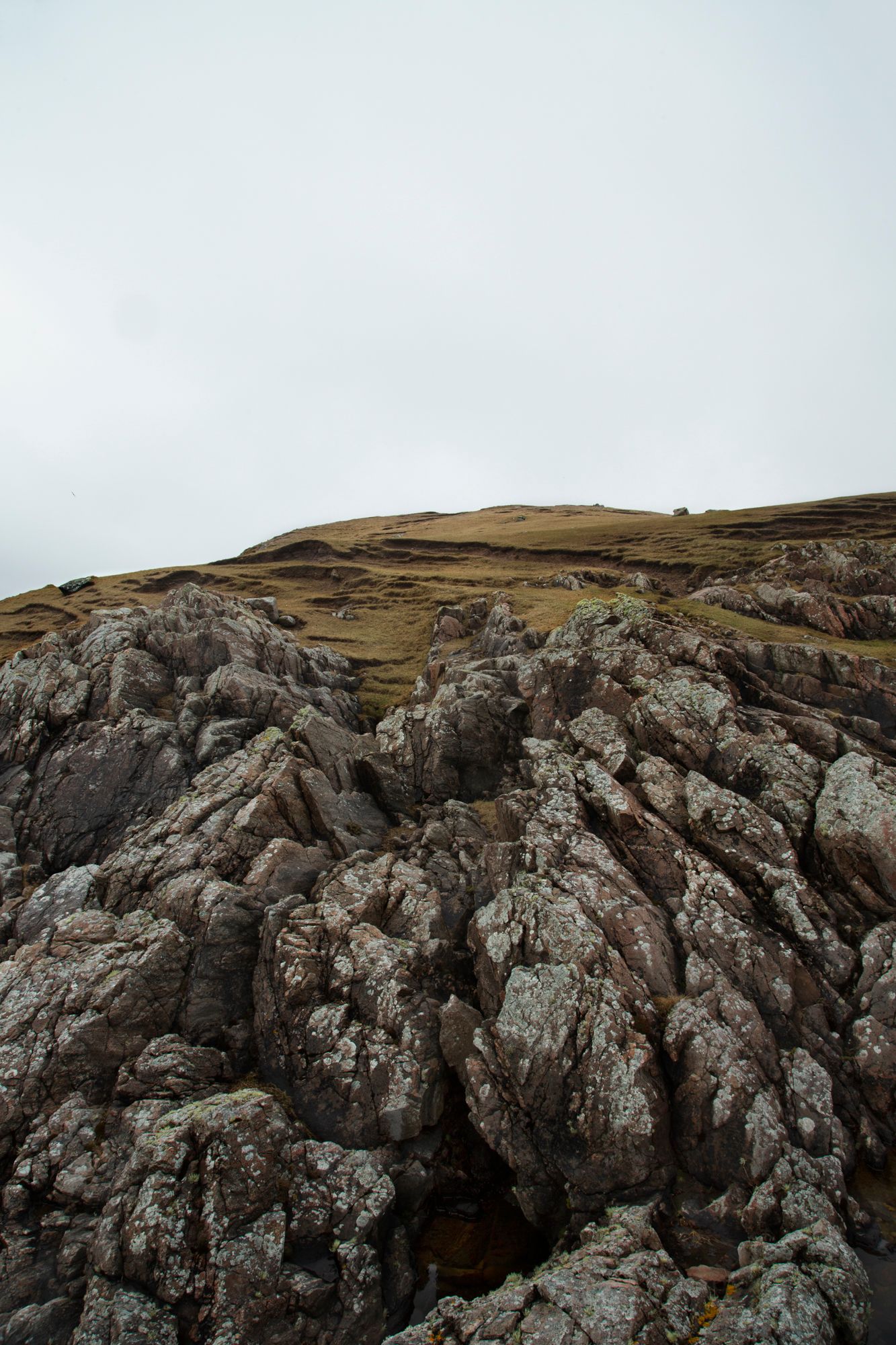 Rugged rocky cliffs and outcrops rise against a cloudy sky in a dramatic coastal landscape.