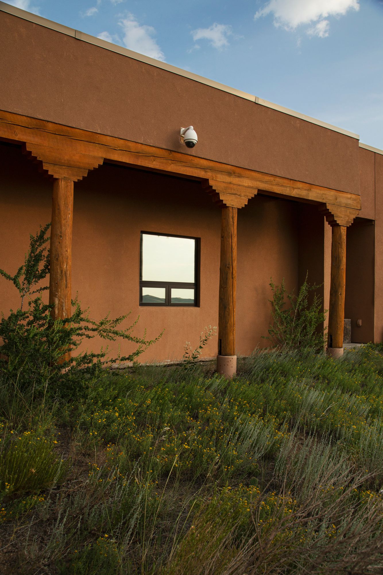 Adobe style buildings with wooden beams and desert landscaping under blue sky with white clouds.