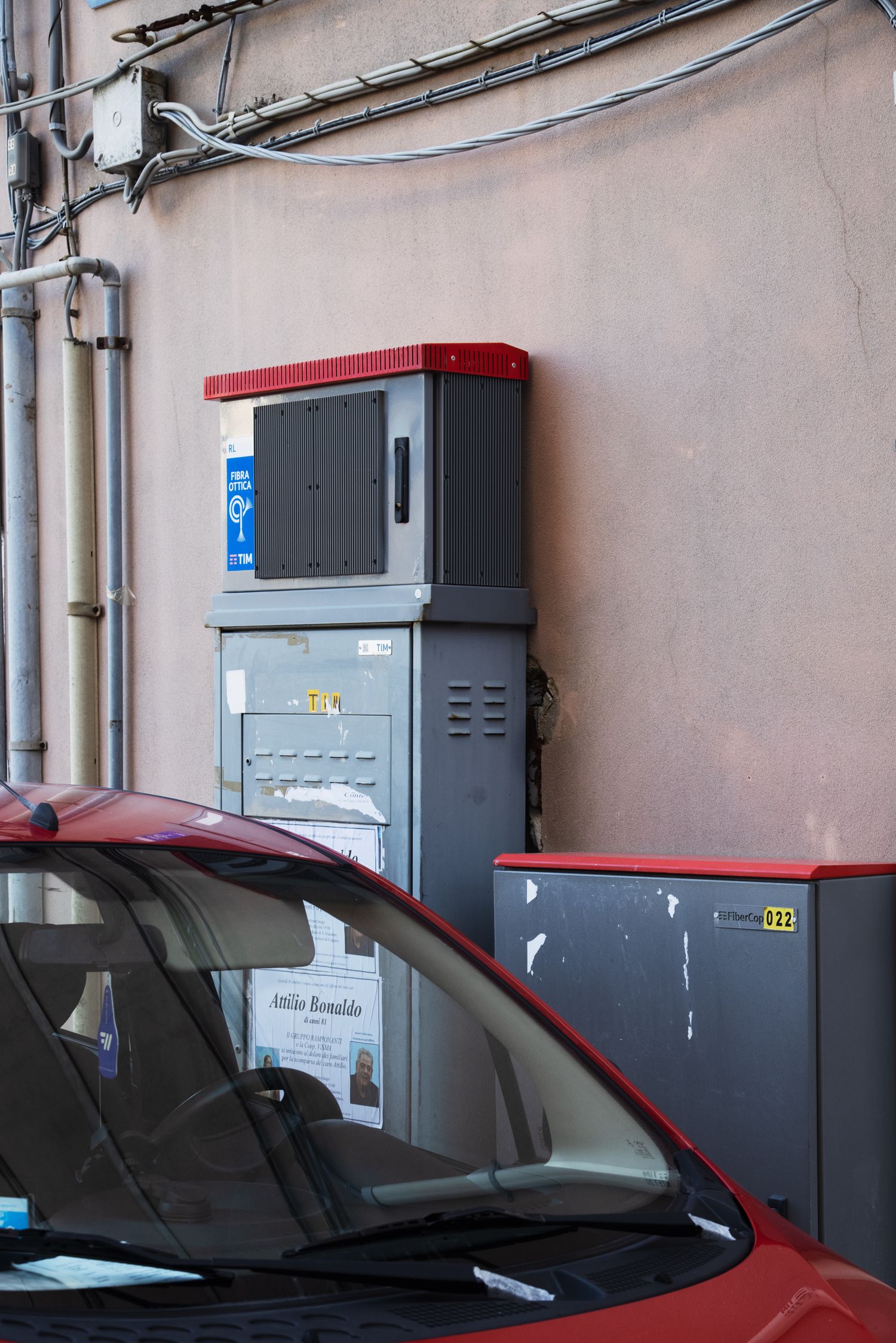 Red car parked near gray electrical utility boxes mounted on a beige wall with overhead wiring.