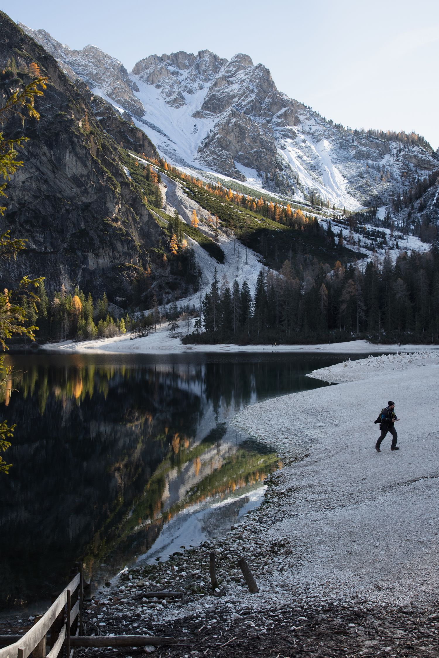 Hiker walks along a reflective alpine lake with snow-capped Dolomite mountains and colorful autumn trees in background.
