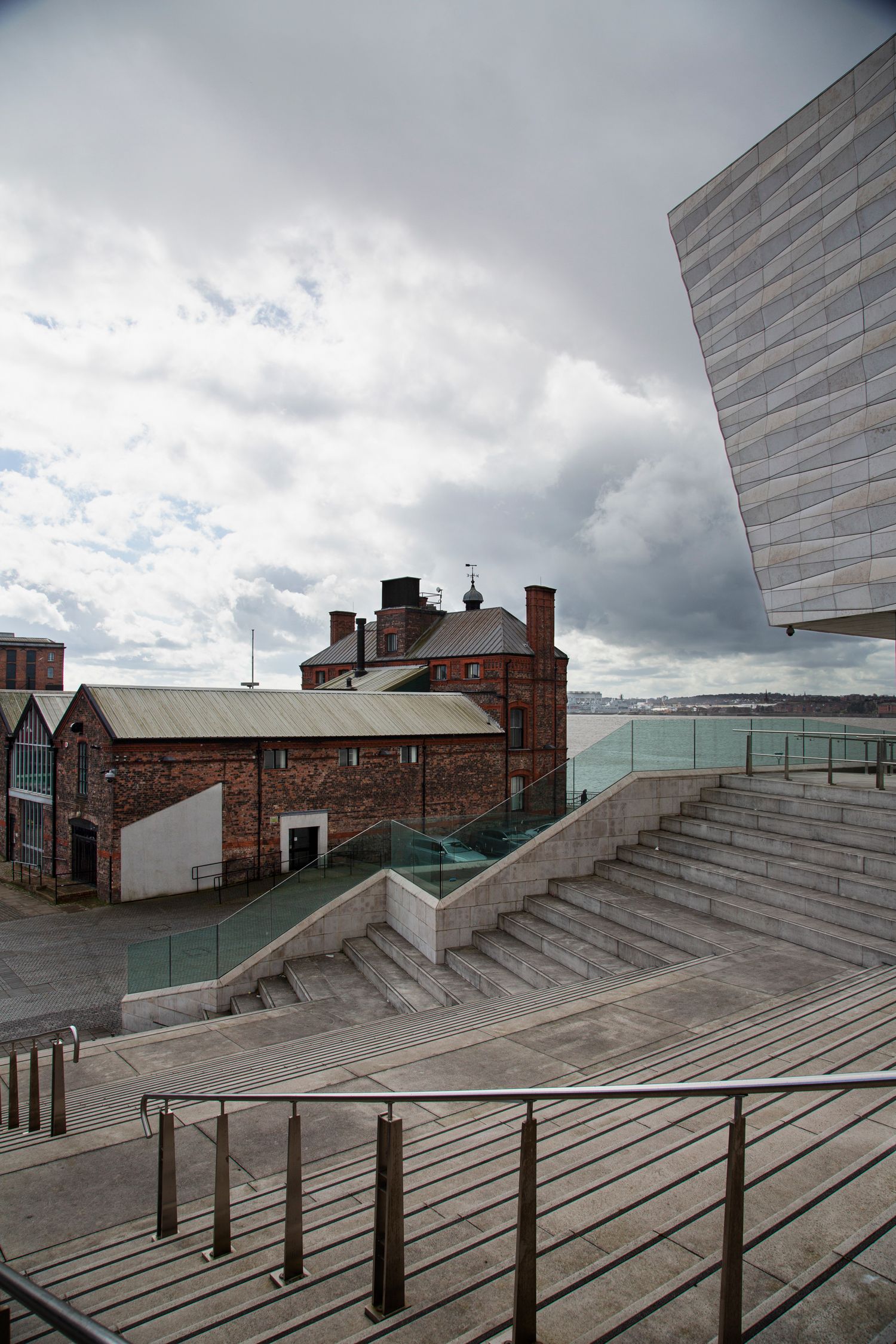 Modern architectural steps with glass railings and brick walls lead up to a building under cloudy skies.