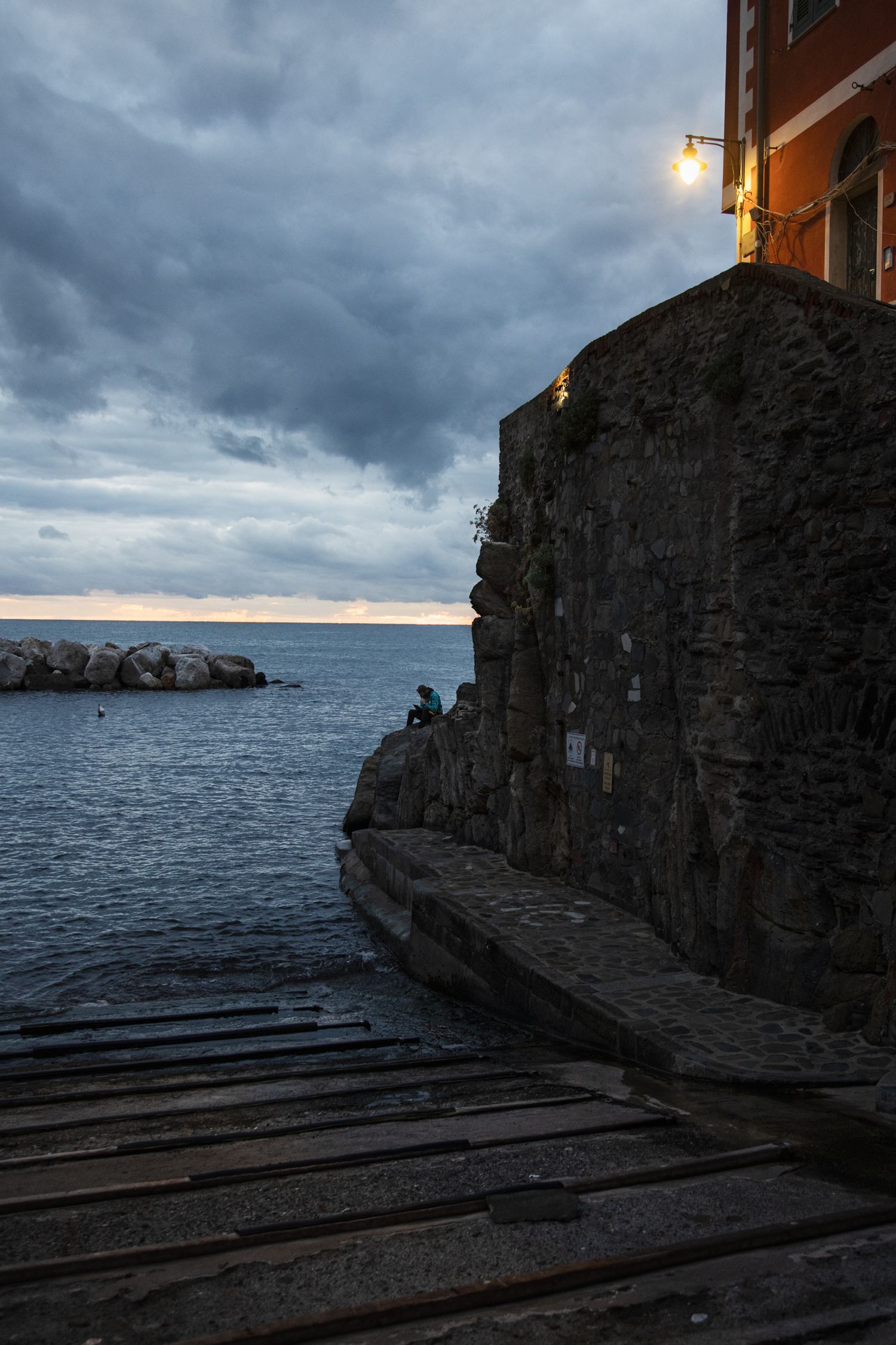 Moody dusk scene at a seaside stone wall with a lone figure sitting by the water under a glowing lantern.