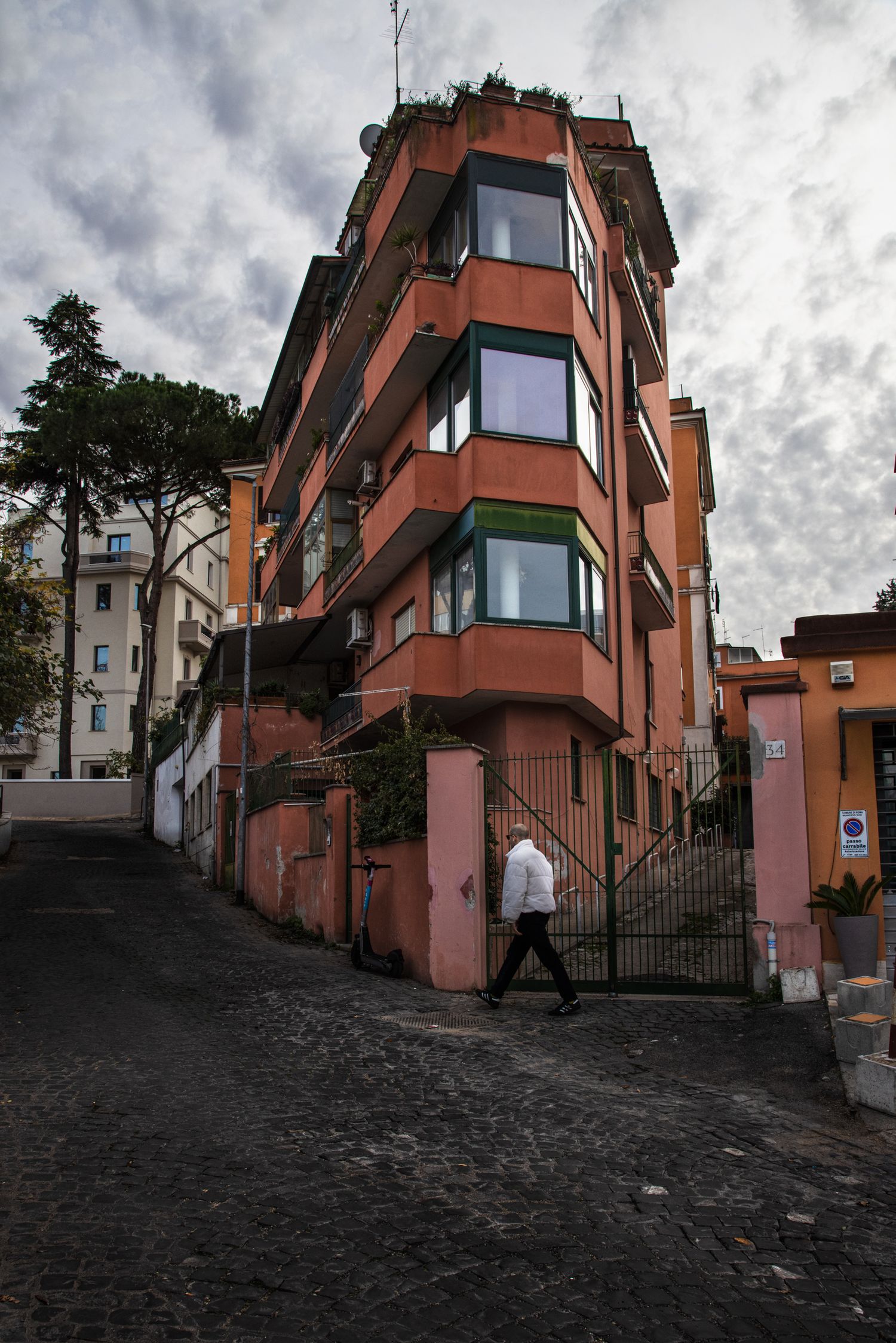 A person walks past a terracotta corner building on a cobblestone street in Rome under a cloudy sky.