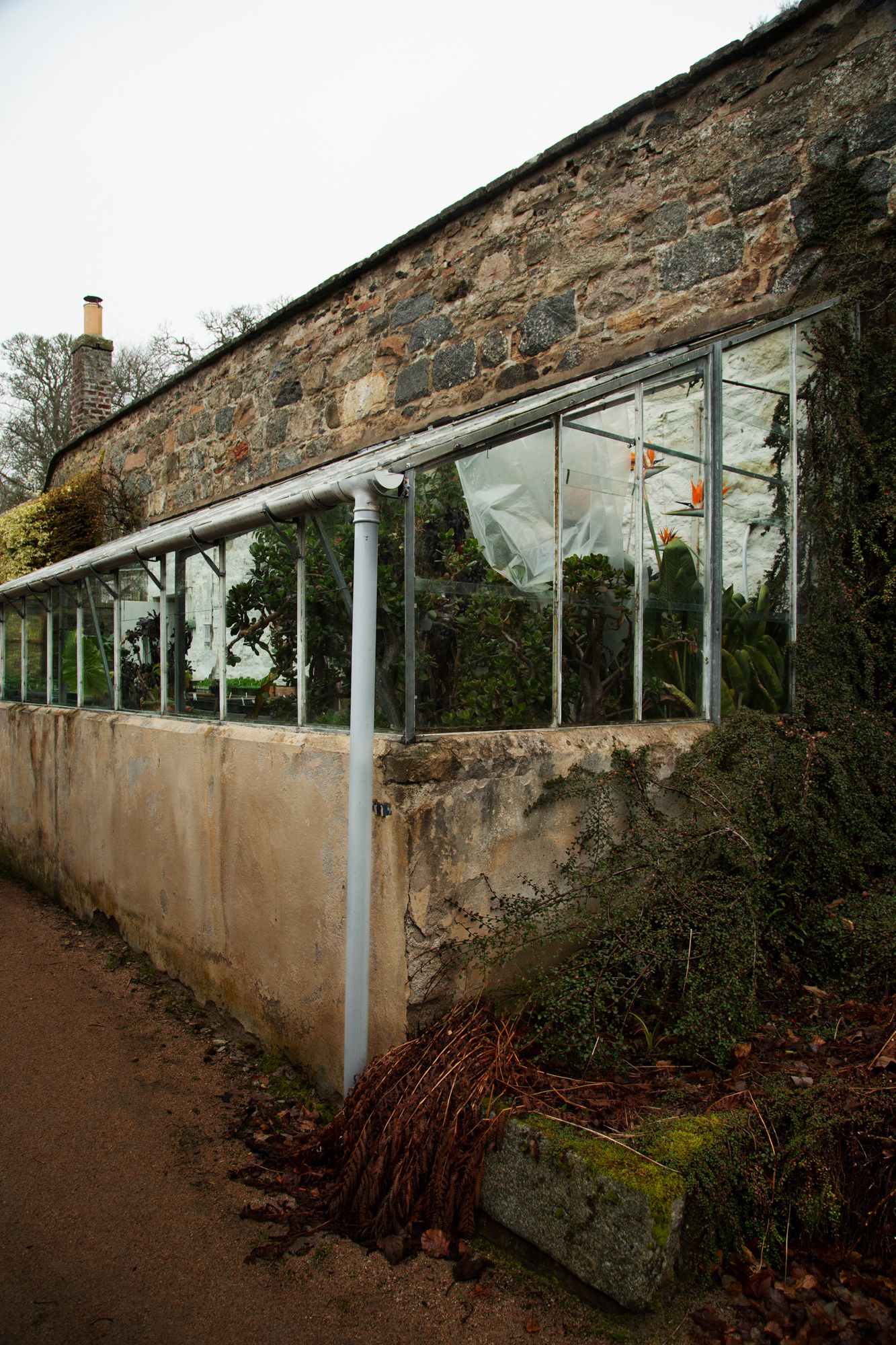 Old stone building with glass greenhouse extension and white support columns along a dirt path.