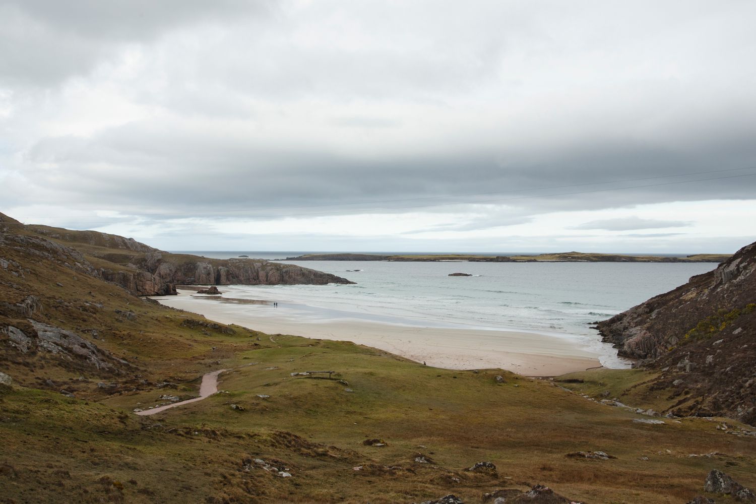 Scenic coastal landscape with dramatic cloudy skies over rugged cliffs and a sandy beach stretching into the distance.