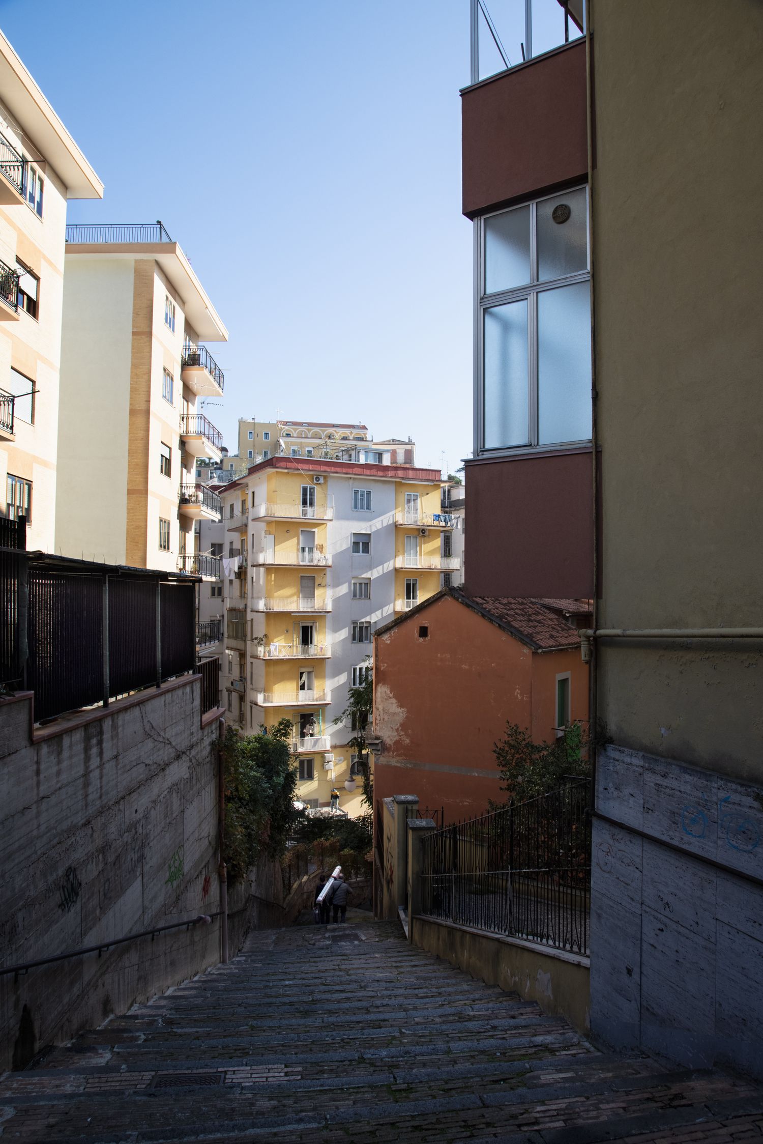 Narrow urban stairway between colorful apartment buildings in an Italian city under a clear blue sky.