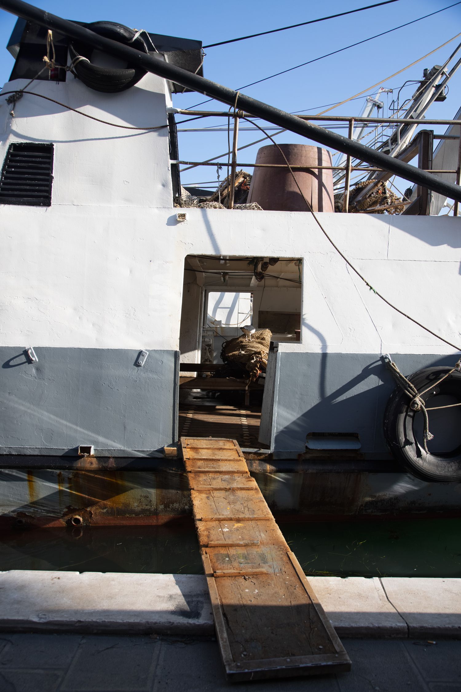 A weathered ship's deck with a wooden gangplank leading to an open hatch, surrounded by industrial equipment and rigging.