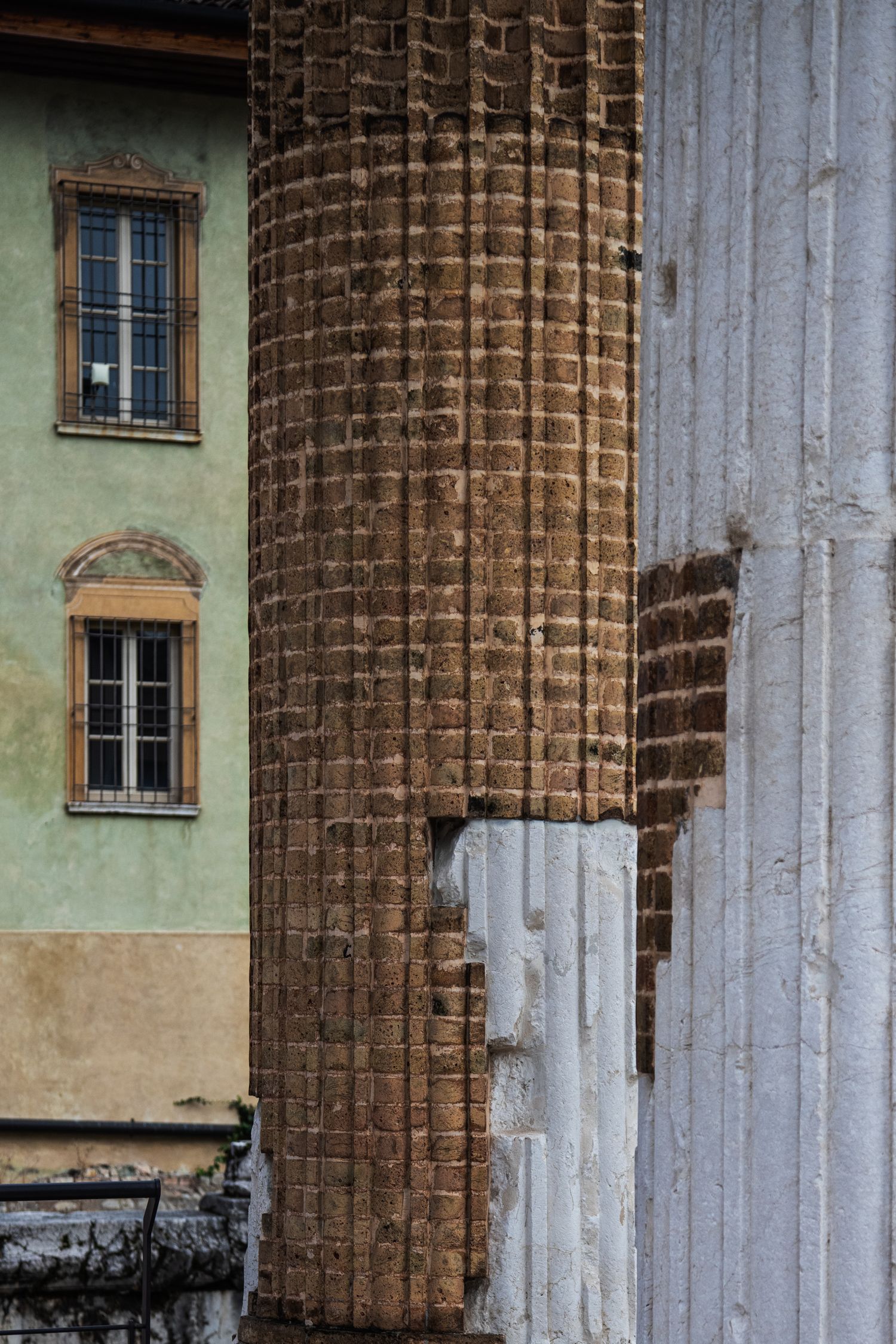 Ancient Roman brick column beside a weathered green building with barred windows in historic Italy.