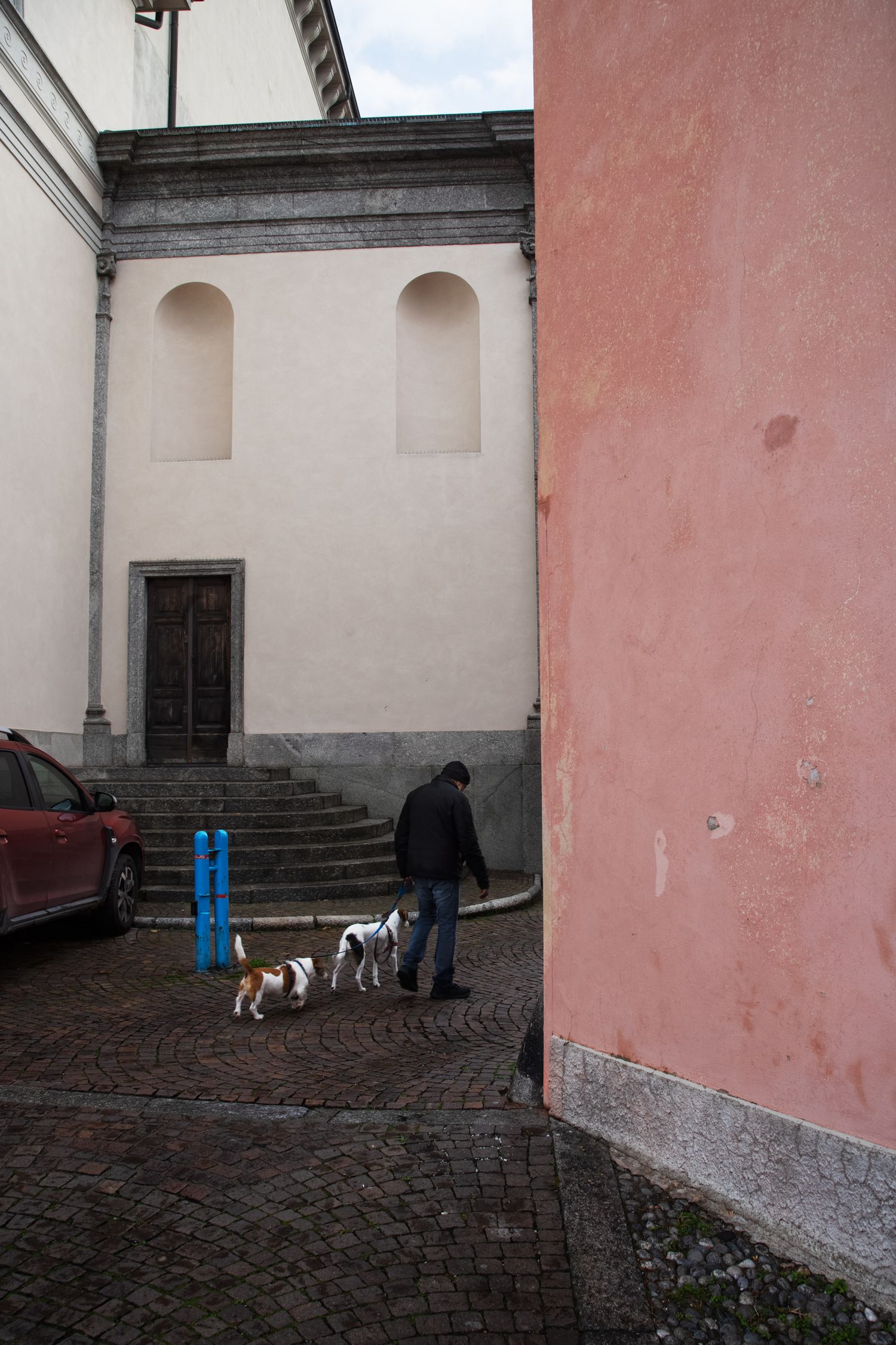 Man walking two small dogs on cobblestone plaza between historic building with arched windows and pink stucco wall.