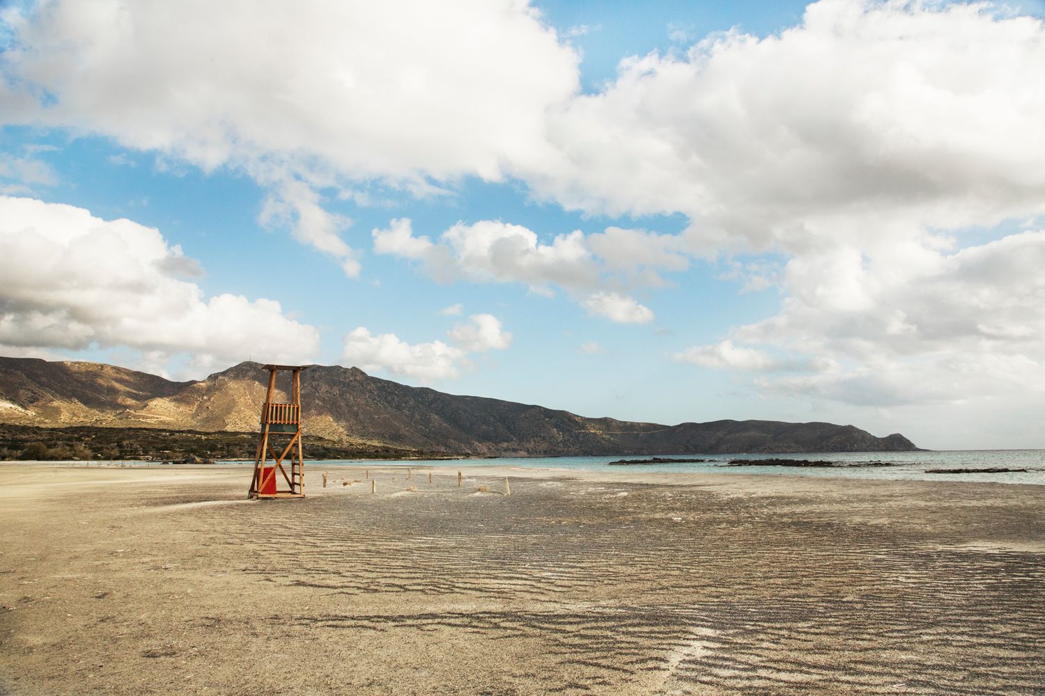 Wide sandy beach stretches along the coastline with mountains and cloudy skies in the background.