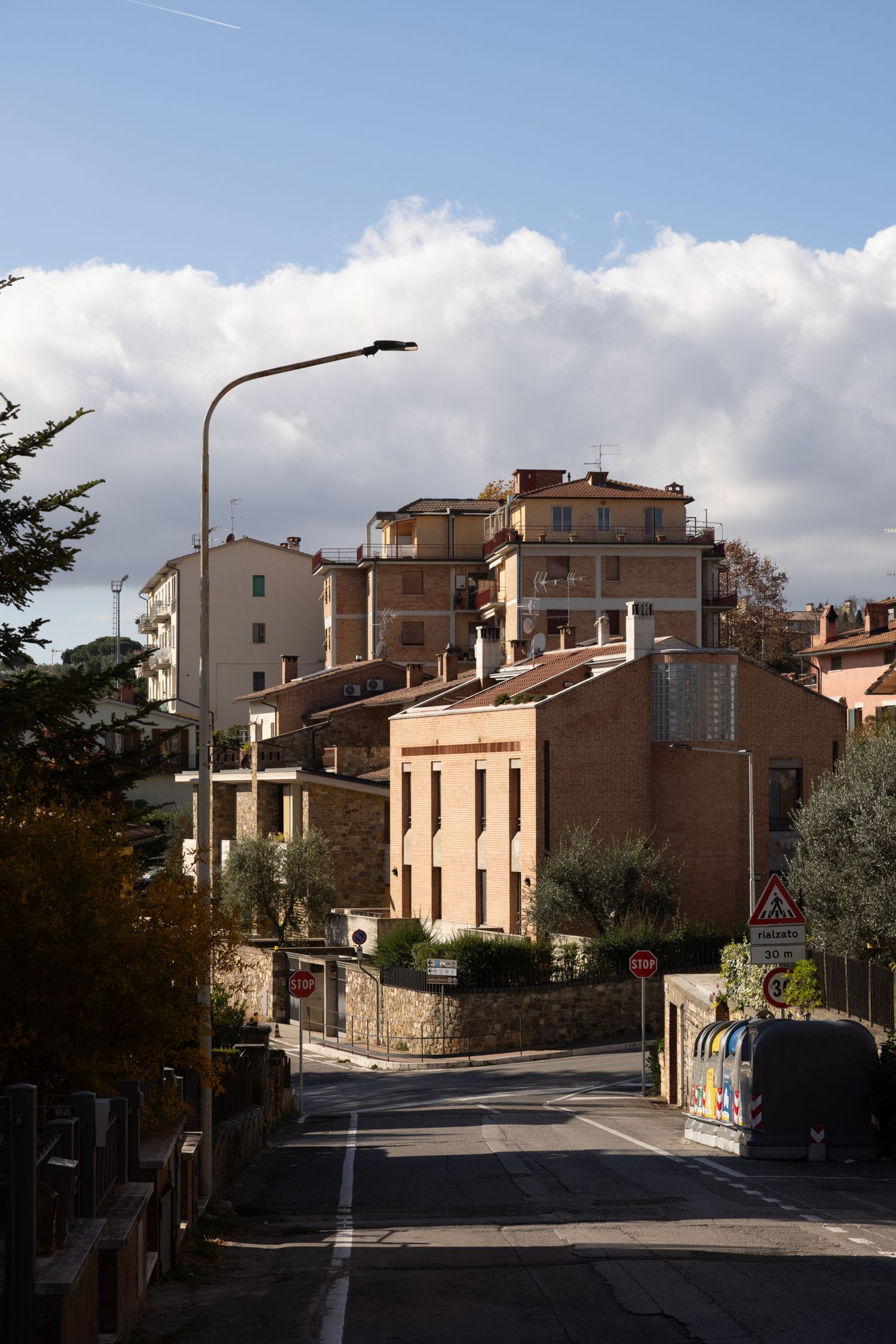 A quiet Italian hillside street with terracotta buildings, a street lamp, and dramatic clouds over a small town.