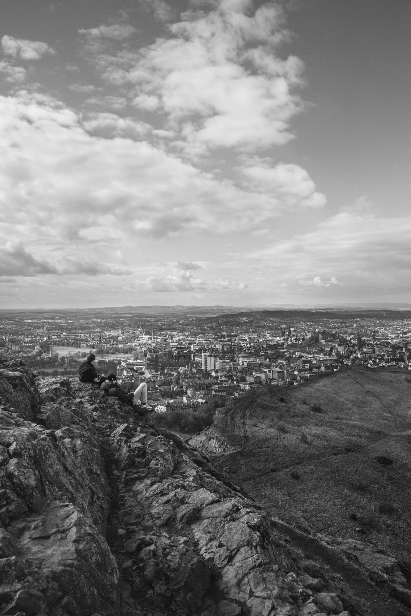 Black and white panoramic view of a coastal city nestled between rocky cliffs and mountains under cloudy skies.