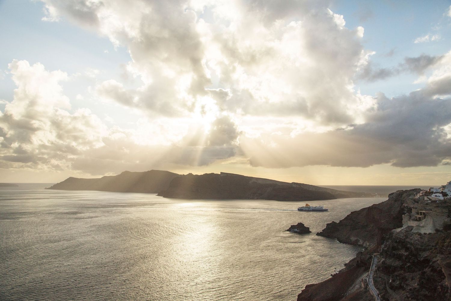 Coastal sunset over ocean with dramatic sun rays breaking through clouds above mountainous shoreline.