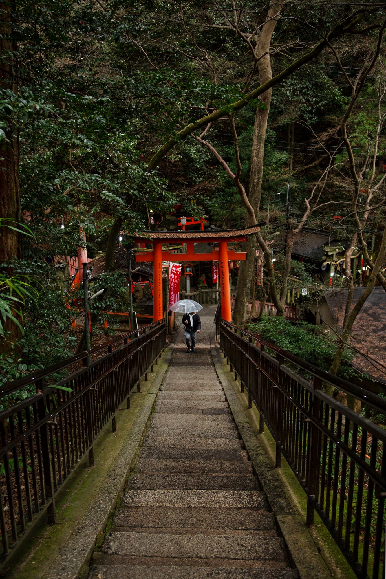 Red Torii gate and bridge path through lush forest with a visitor holding an umbrella.