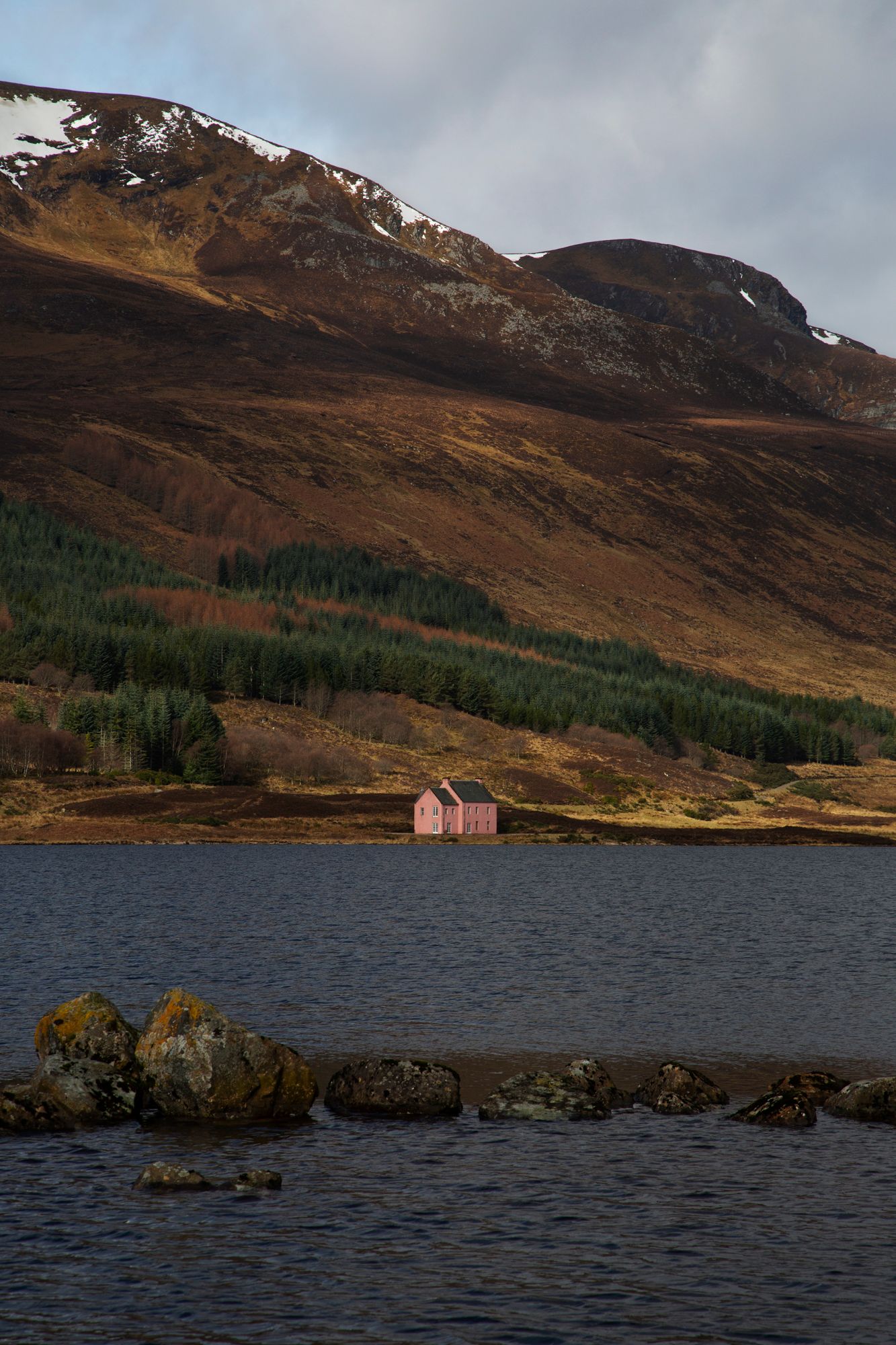 A pink cottage sits on the shores of a Scottish loch with rugged mountains and autumn foliage in the background.