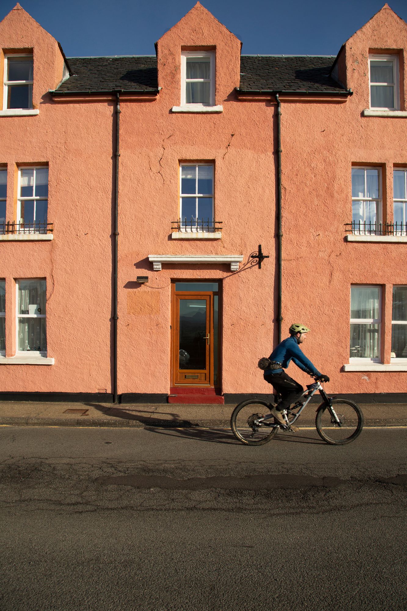 A cyclist rides past a coral-colored building with dormer windows on a quiet street.