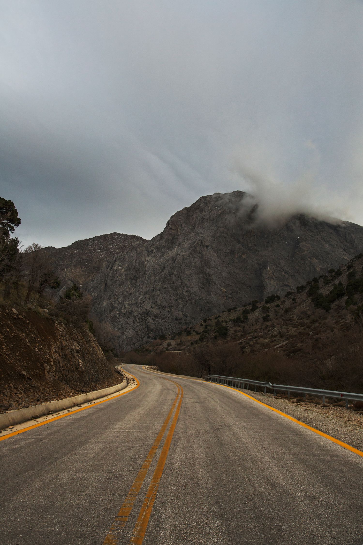 Winding mountain road with yellow center lines cutting through rocky terrain under cloudy skies.