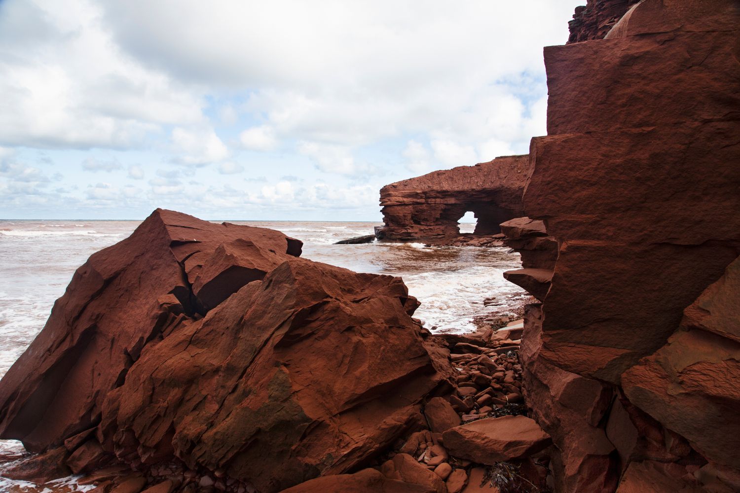 Dramatic red rock formations and cliffs overlook a turbulent ocean coastline.