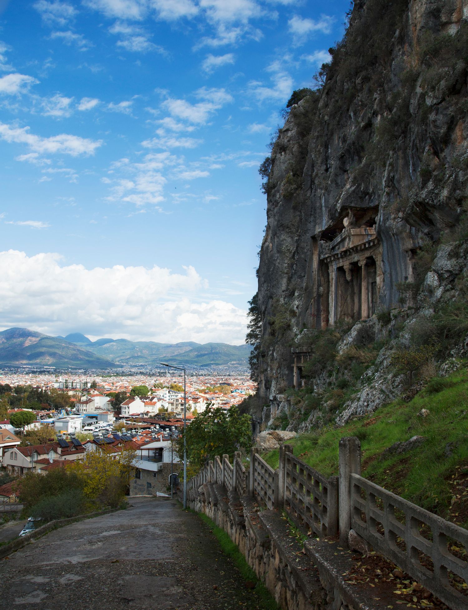 Stone pathway along a cliff edge overlooks a scenic cityscape with mountains in the background.
