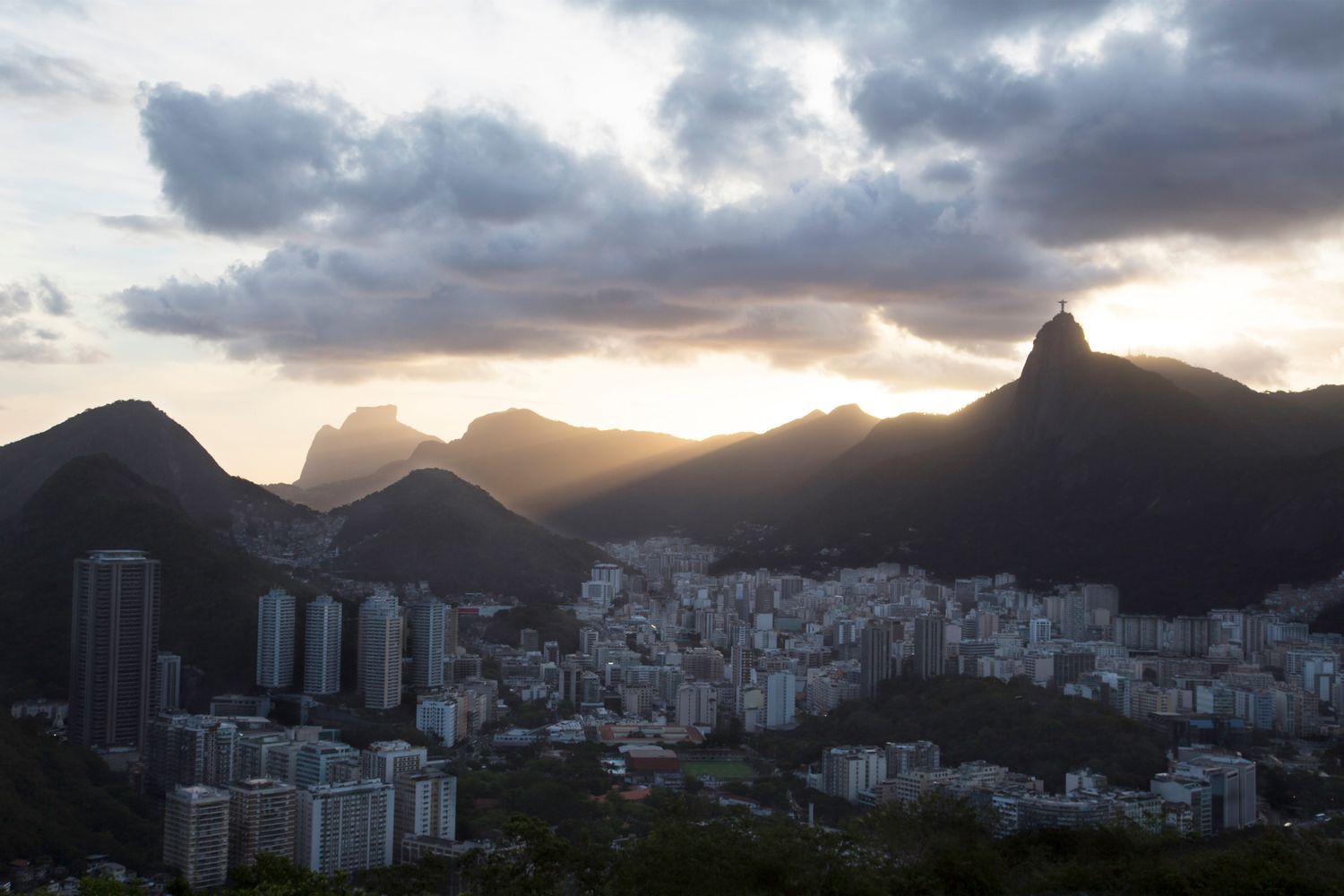 Panoramic cityscape at sunset with mountains and high-rise buildings silhouetted against dramatic sky.