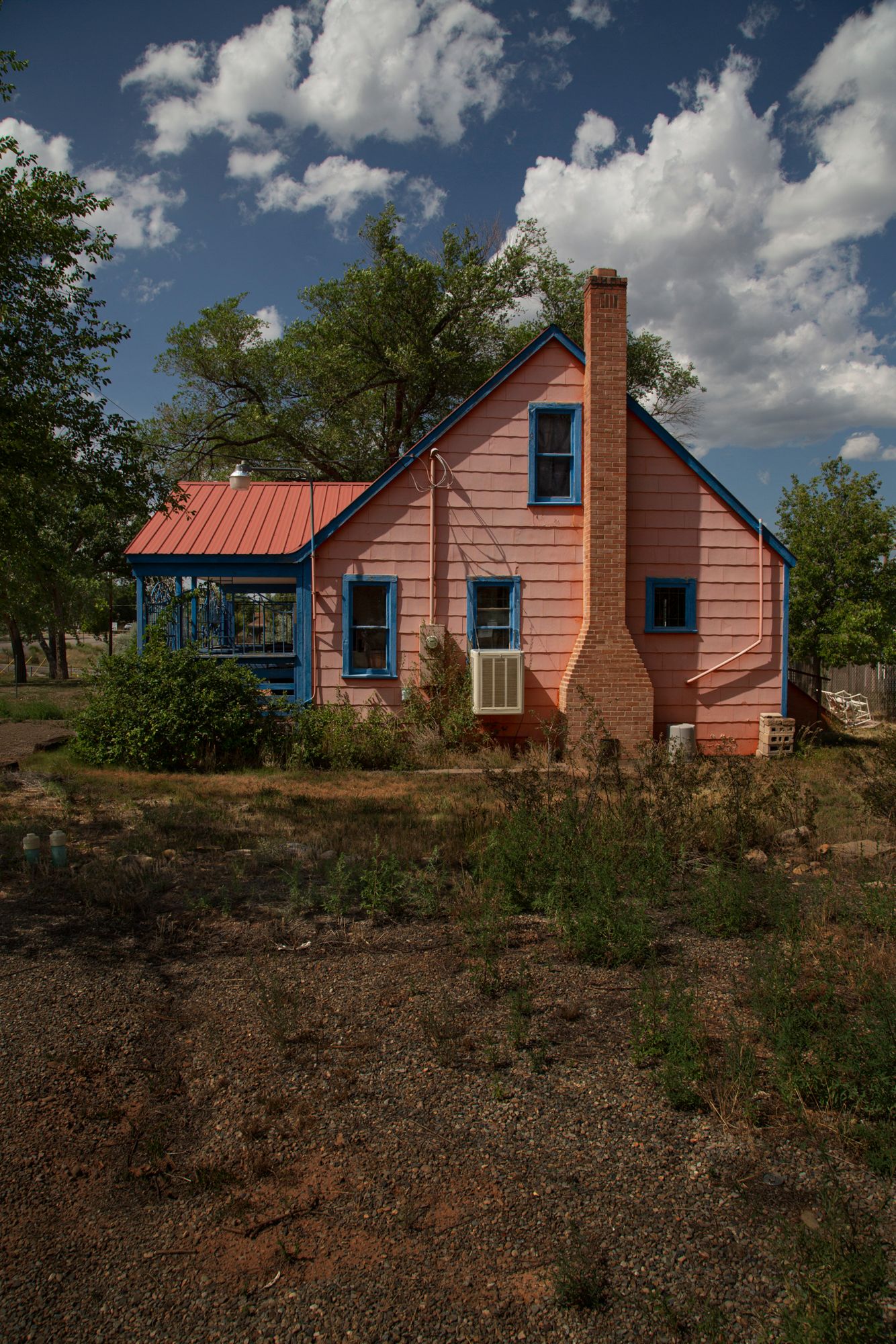 A rustic red wooden house with blue trim and a brick chimney sits atop a hillside under puffy white clouds.