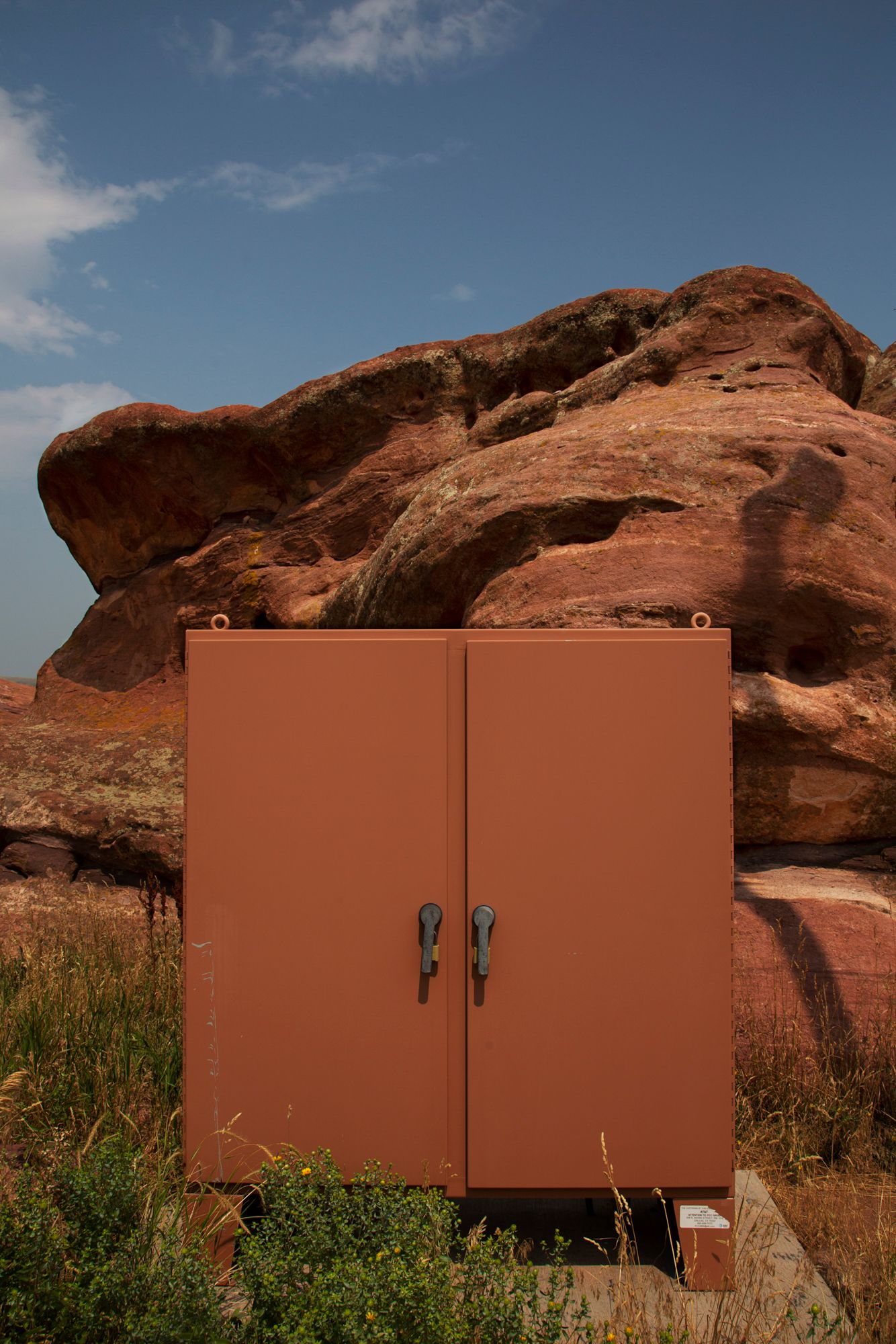 A brown metal utility cabinet stands against red rock formations in a desert landscape under blue skies.
