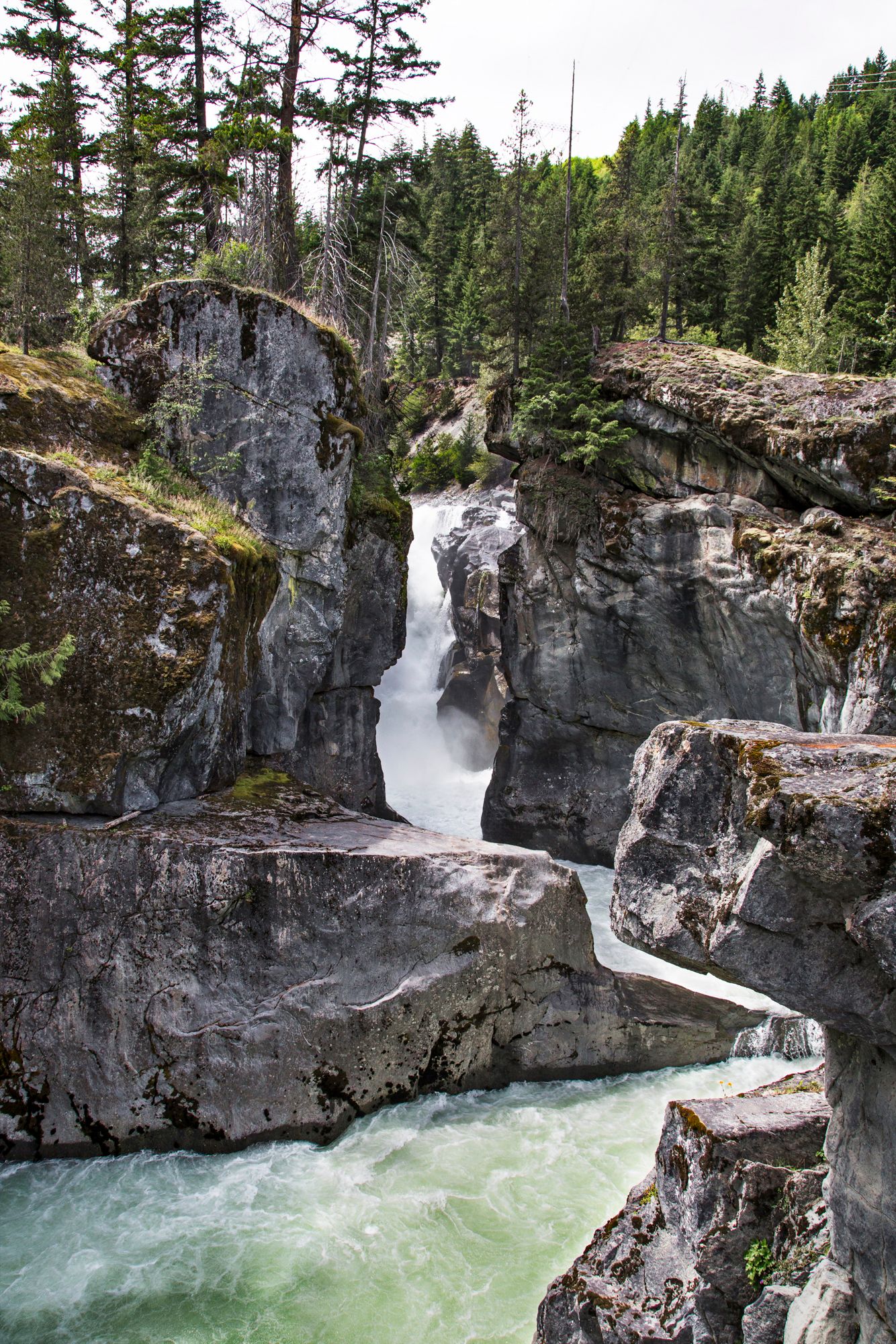 Dramatic waterfall cuts through rocky gorge with turquoise water flowing beneath evergreen forest.
