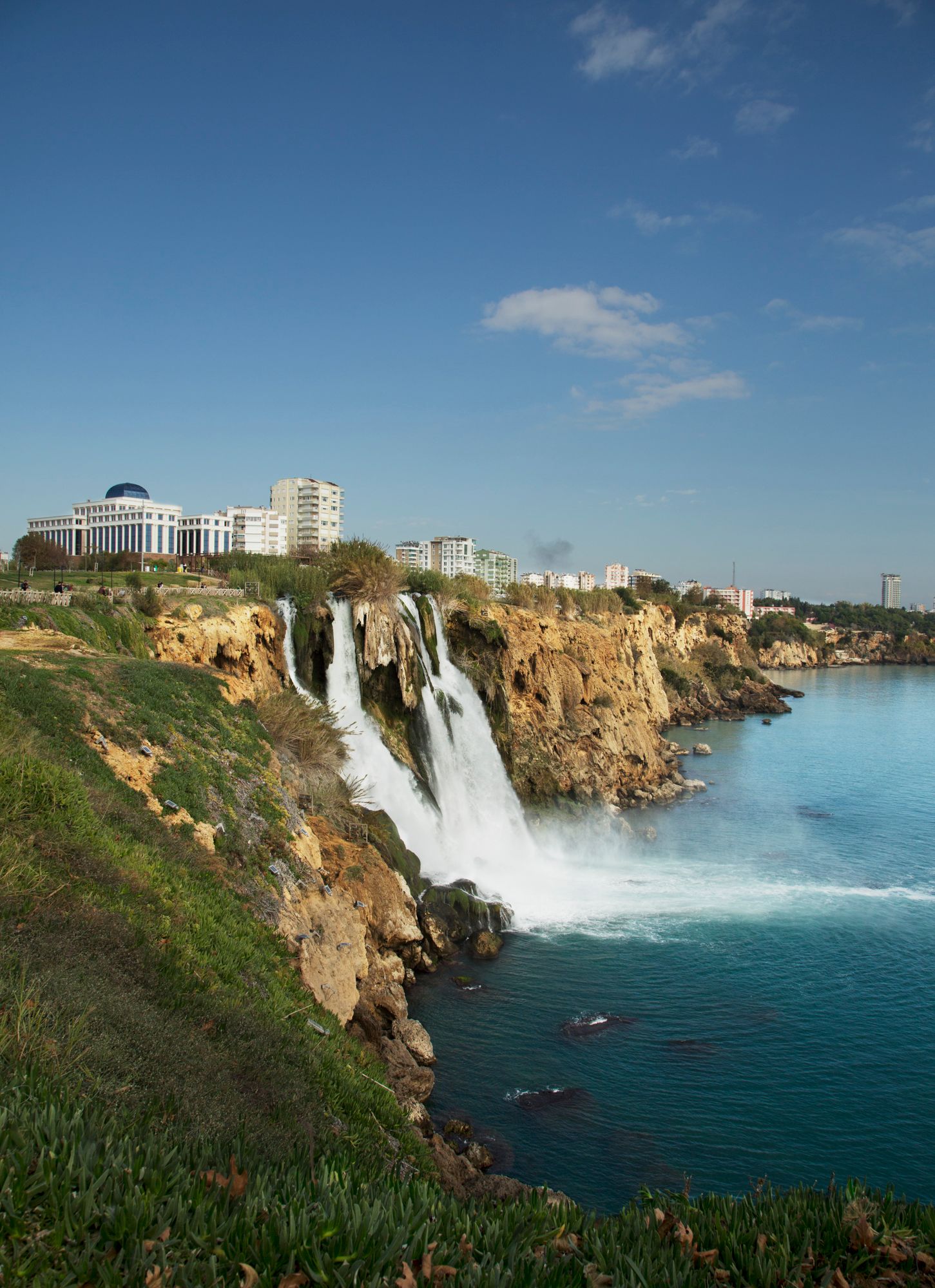 Dramatic waterfall cascades down rocky cliffs into turquoise Mediterranean waters near a coastal city.