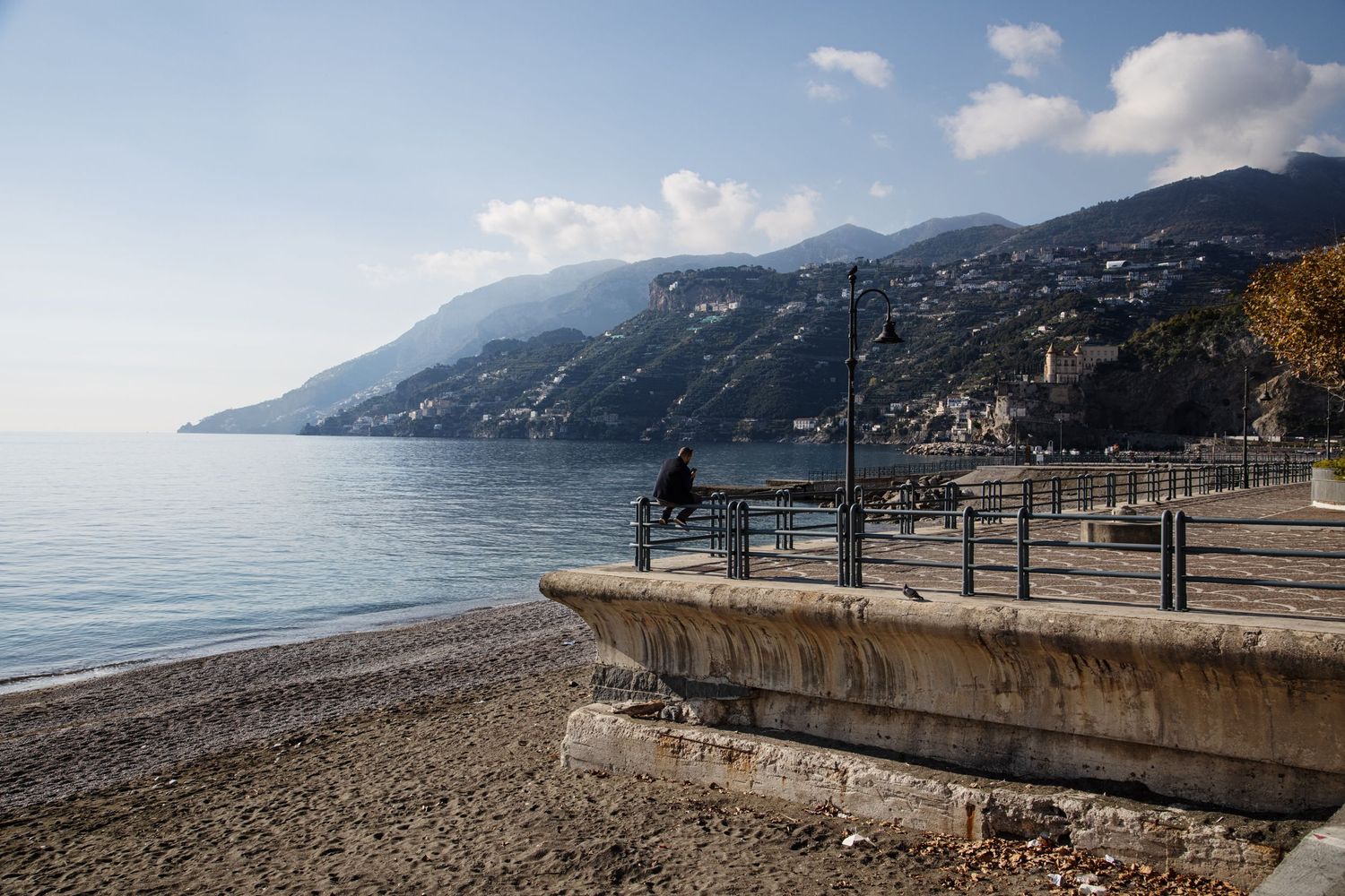 Person sitting on a seaside promenade wall overlooking calm blue waters and mountain coastline on a sunny day.