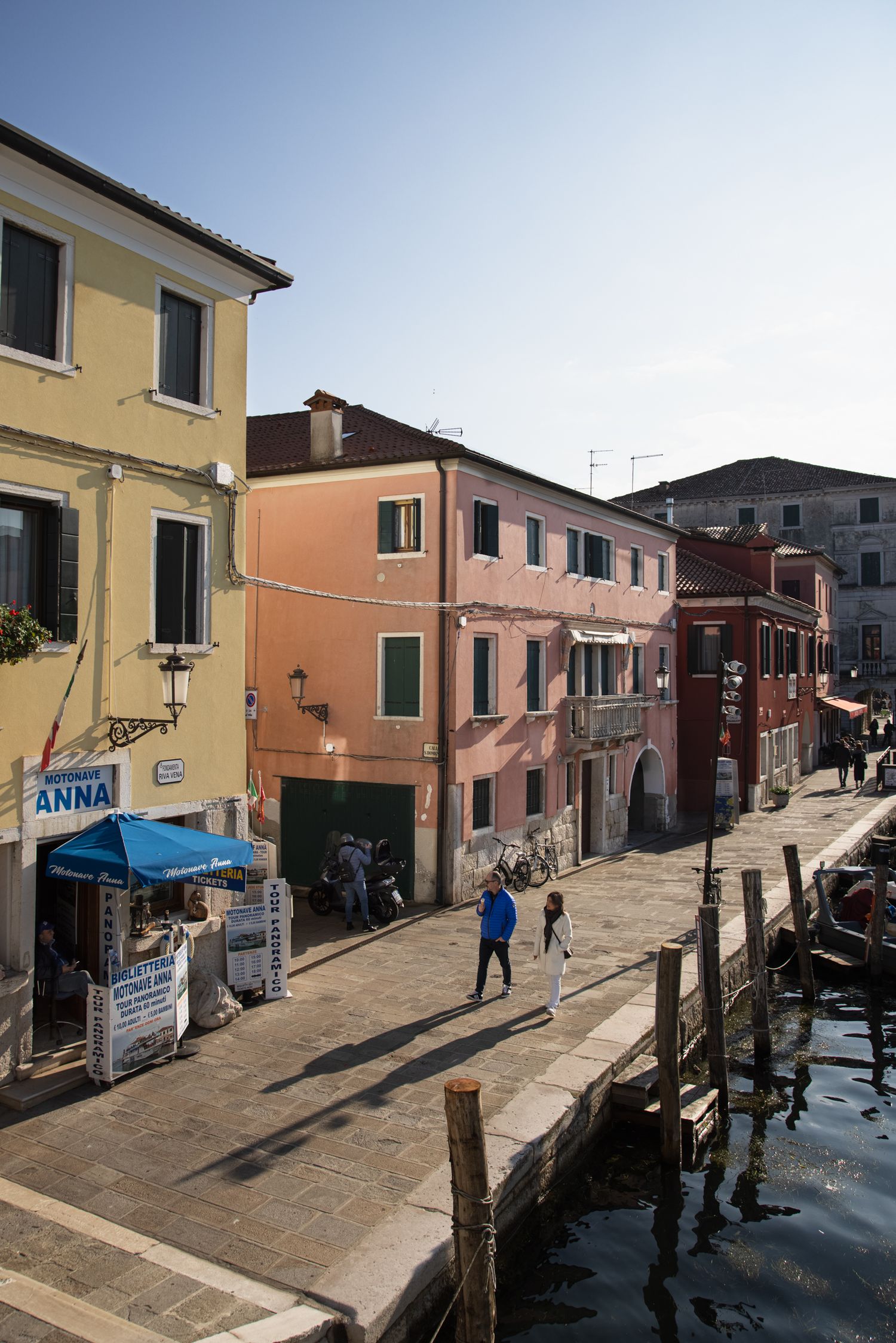 Colorful buildings line a sunny Venice canal walkway with pedestrians strolling along the waterfront promenade.