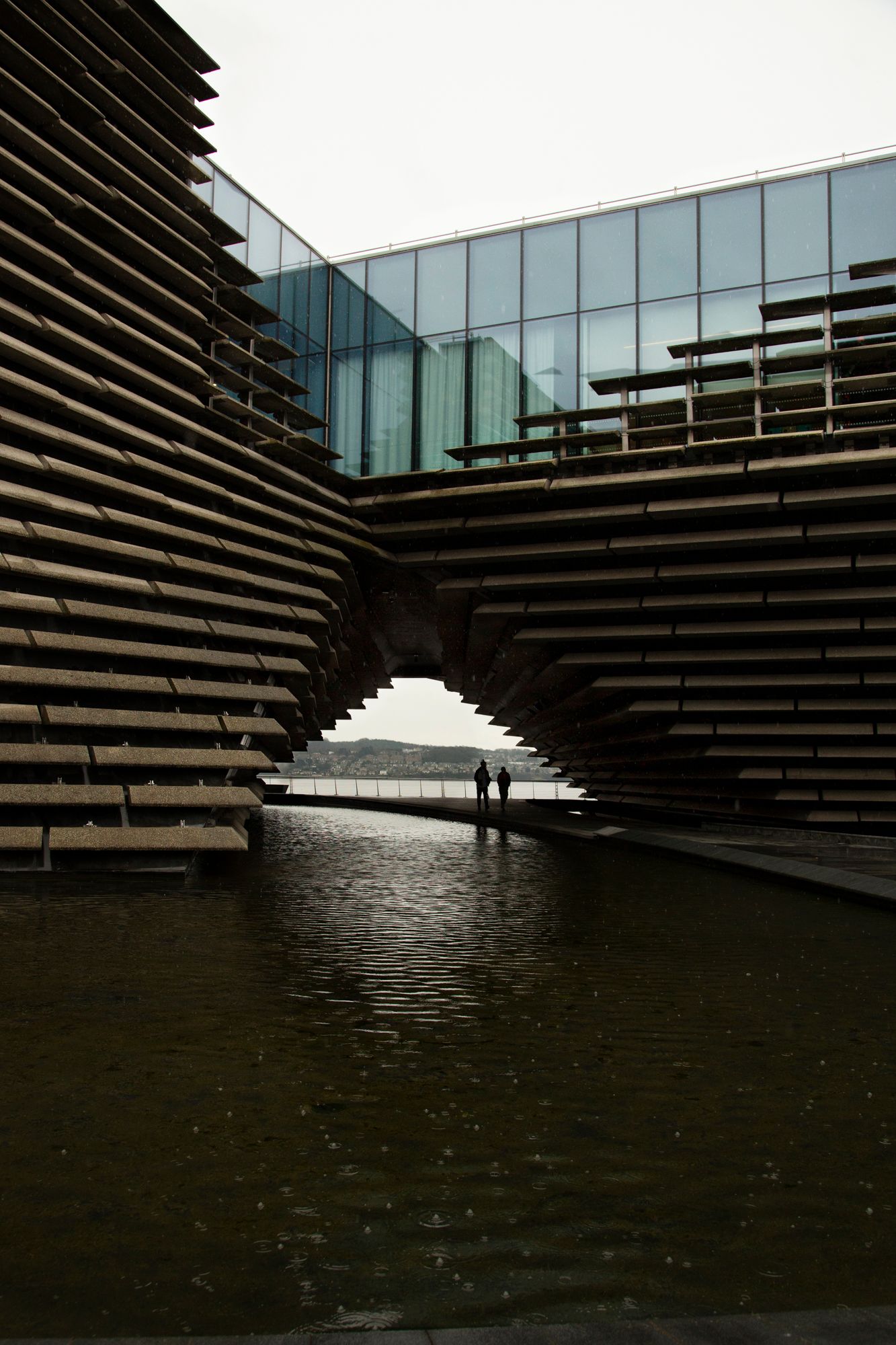 Modern architectural building with layered wooden slats reflects in dark water pool at V&A Dundee museum in Scotland.