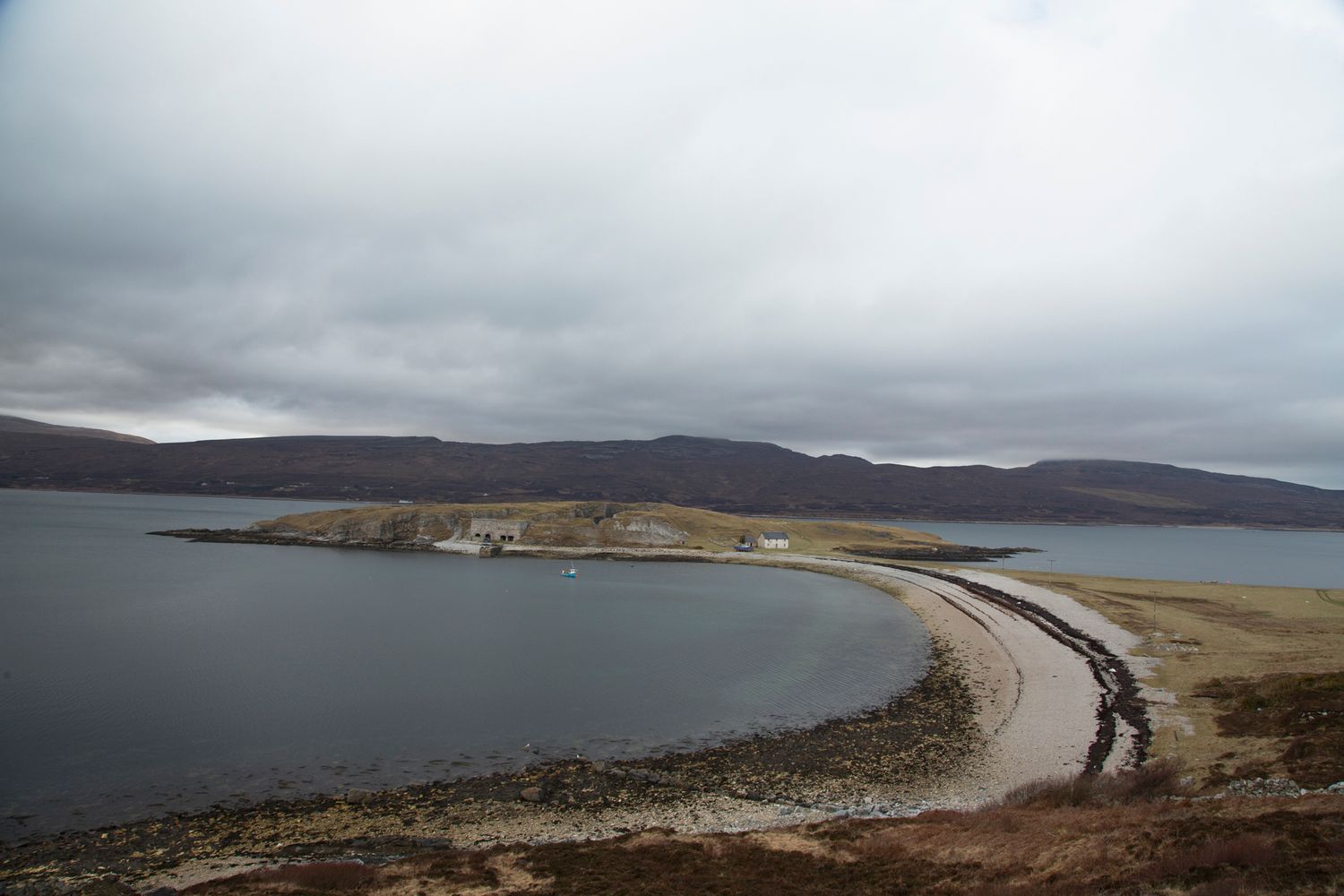 A curved causeway road connects to a rocky island under moody gray skies over calm waters in Scotland.