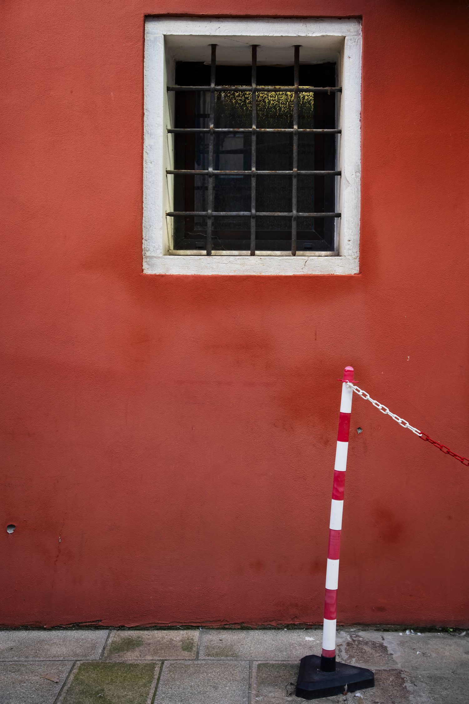 Red wall with barred window and red-white striped barrier pole on a cobblestone sidewalk.