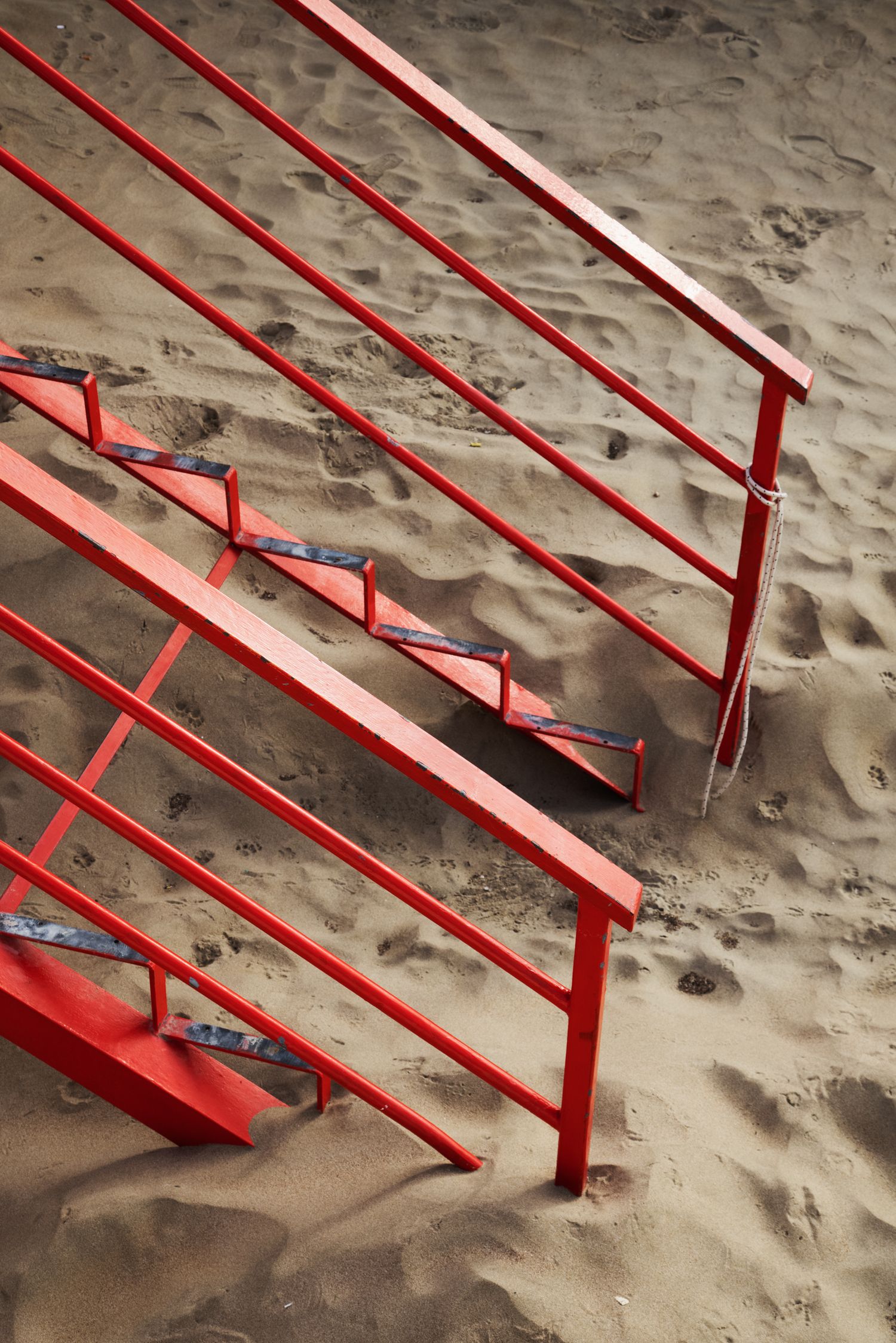 Red metal staircase with railings on sandy beach, vibrant contrast against tan sand with visible footprints.