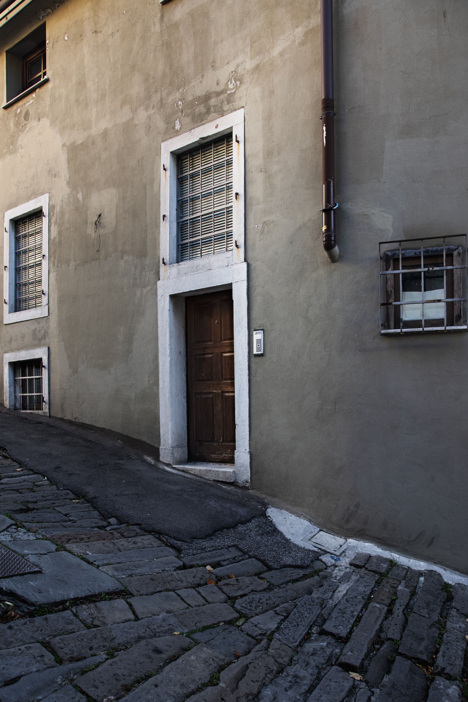 Narrow cobblestone alley with aged gray building, barred windows, and wooden door in historic European town.
