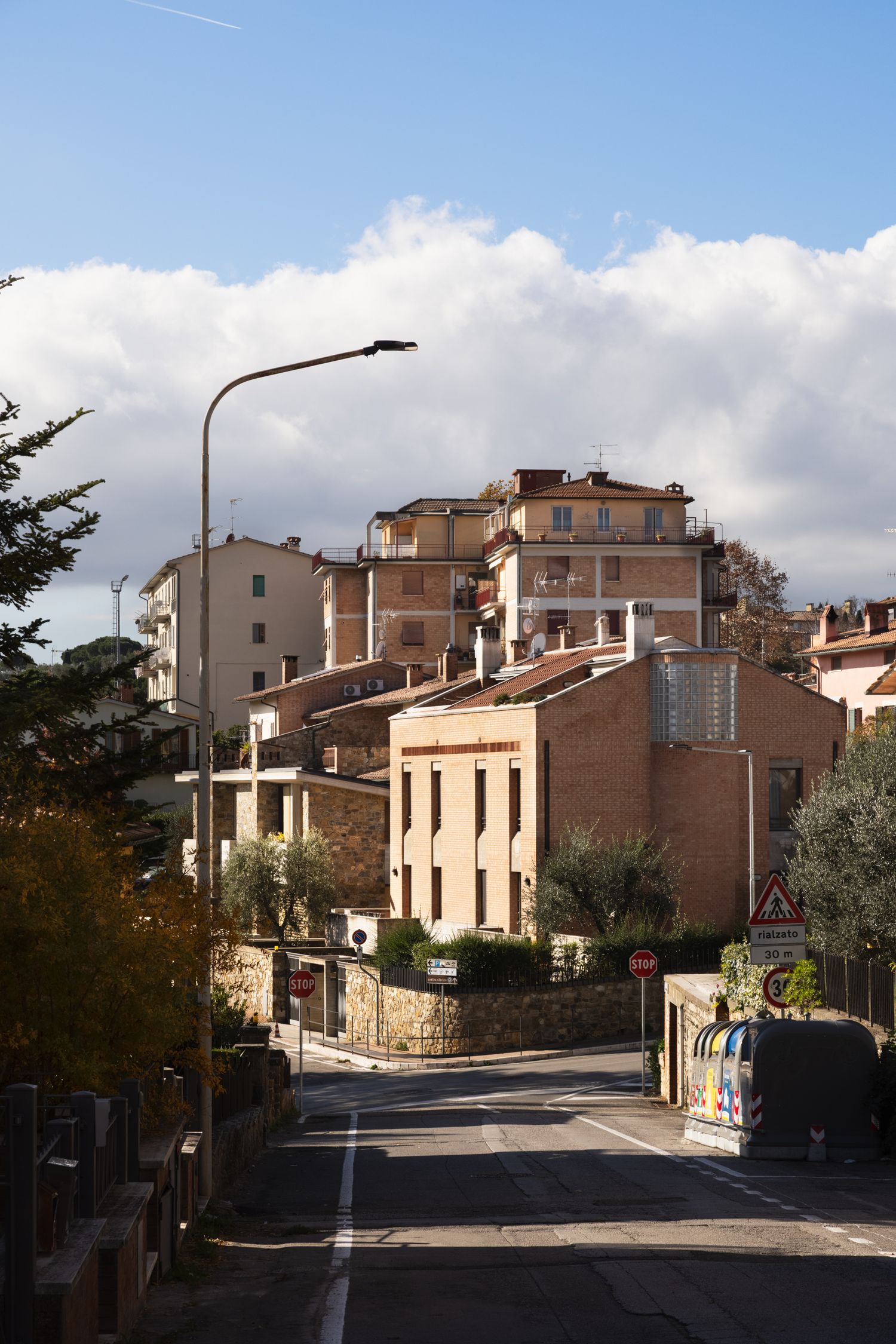 Sunlit Italian hillside town with terracotta buildings, a quiet road, street lamp, and dramatic clouds in a blue sky.