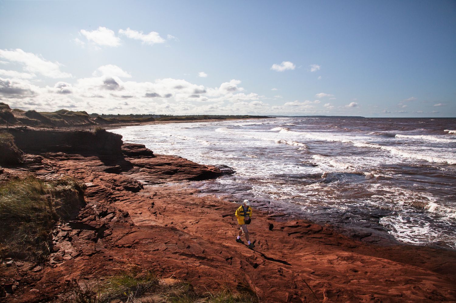 Waves break against red rocky shoreline under scattered clouds and bright sunlight.