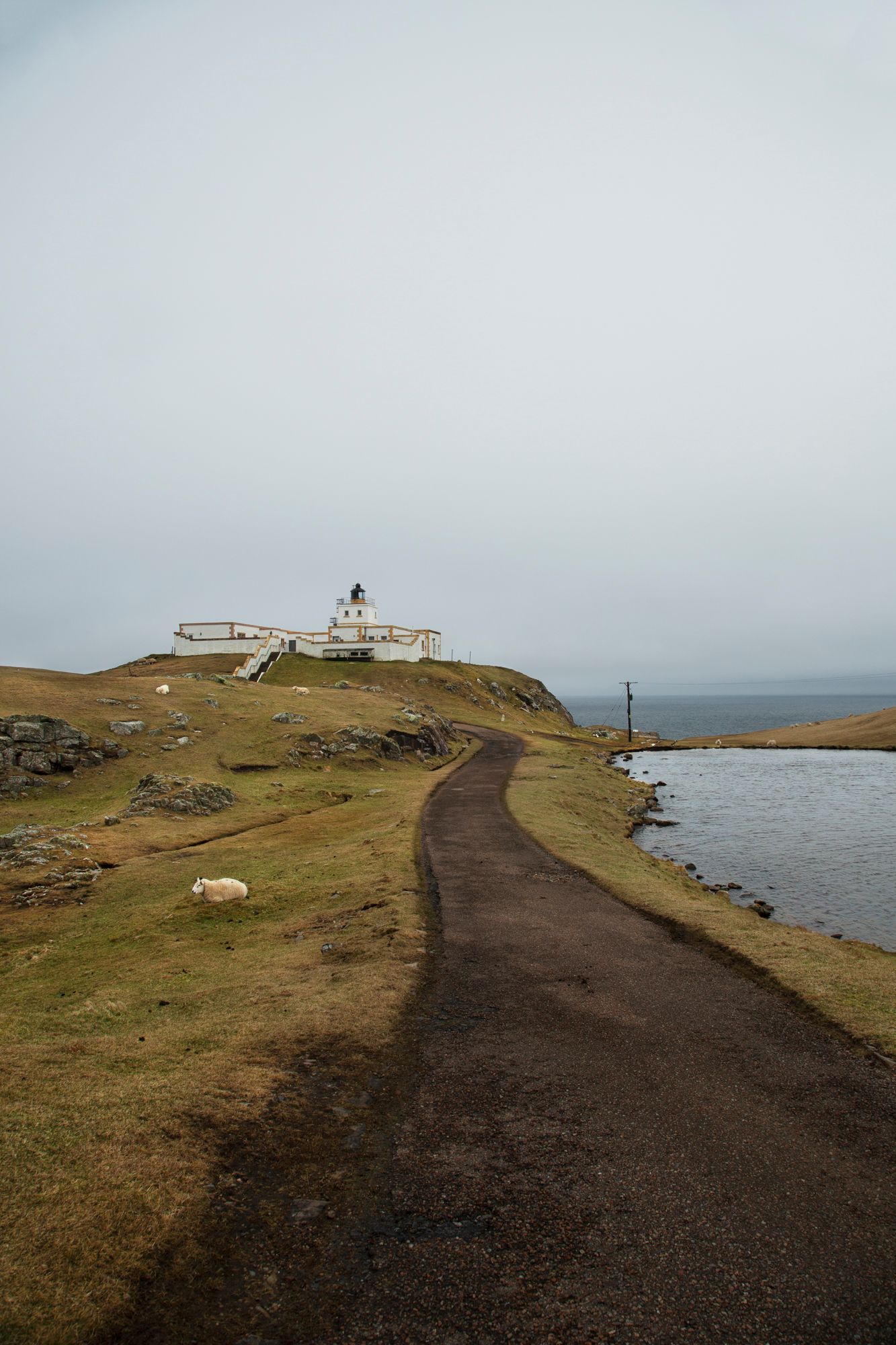 A winding paved path leads to a lighthouse on a rugged coastal cliff overlooking the ocean on a cloudy day.