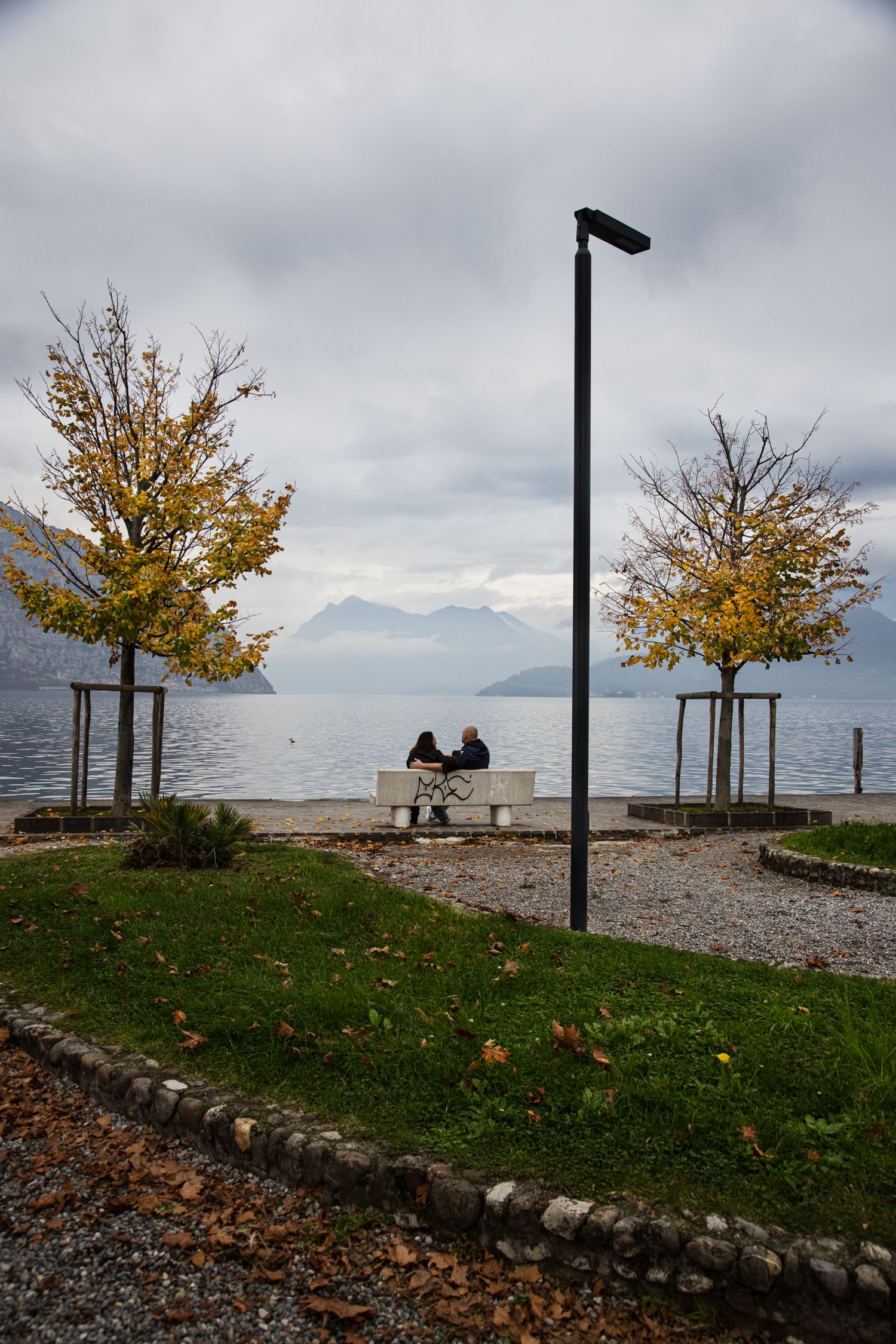 Two people sit on a bench by a lake in autumn, framed by yellow trees, mountains, and a street lamp under cloudy skies.
