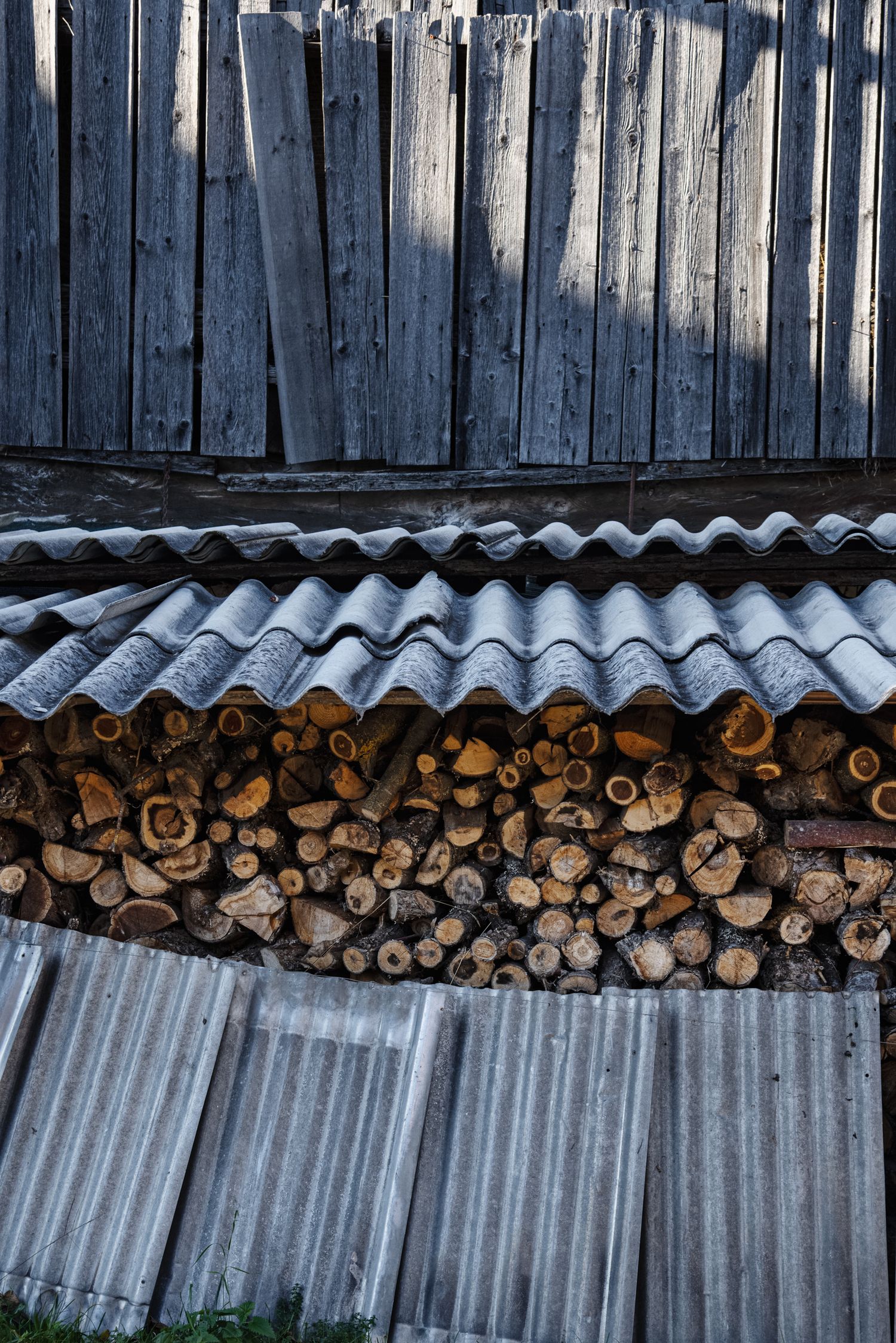 Stacked firewood logs stored under a corrugated metal roof shelter against a weathered wooden fence.