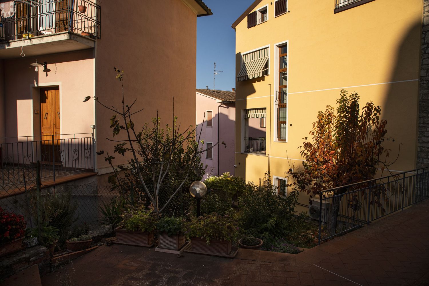 Colorful Italian courtyard with lush green plants, yellow and pink building facades, and iron railings in warm sunlight.