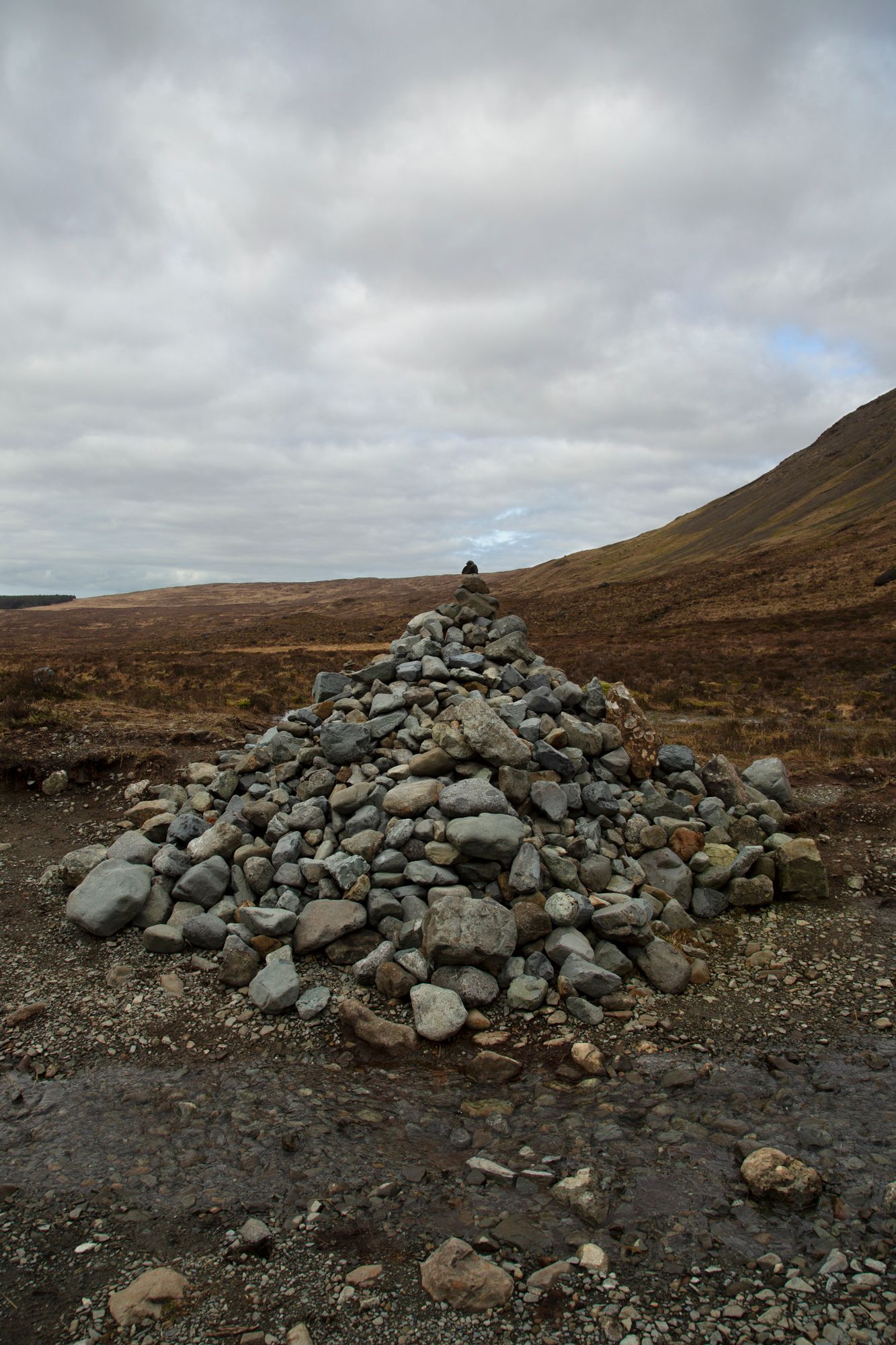 A stone cairn stands against a cloudy sky on a barren hillside in the Scottish Highlands.