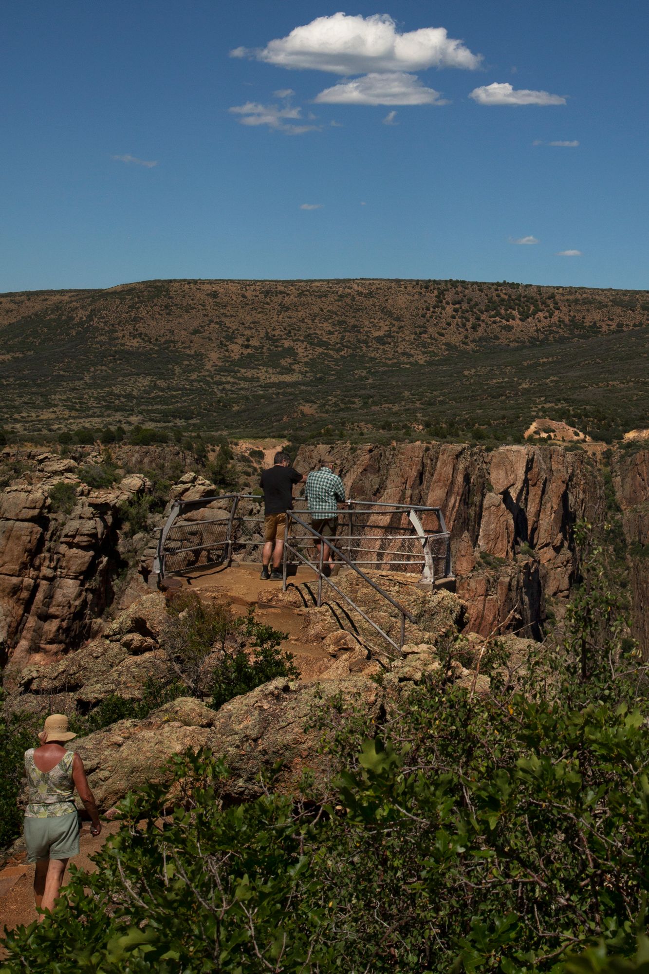 Visitors stand at a scenic overlook with metal railings above rugged canyon cliffs under a bright blue desert sky.