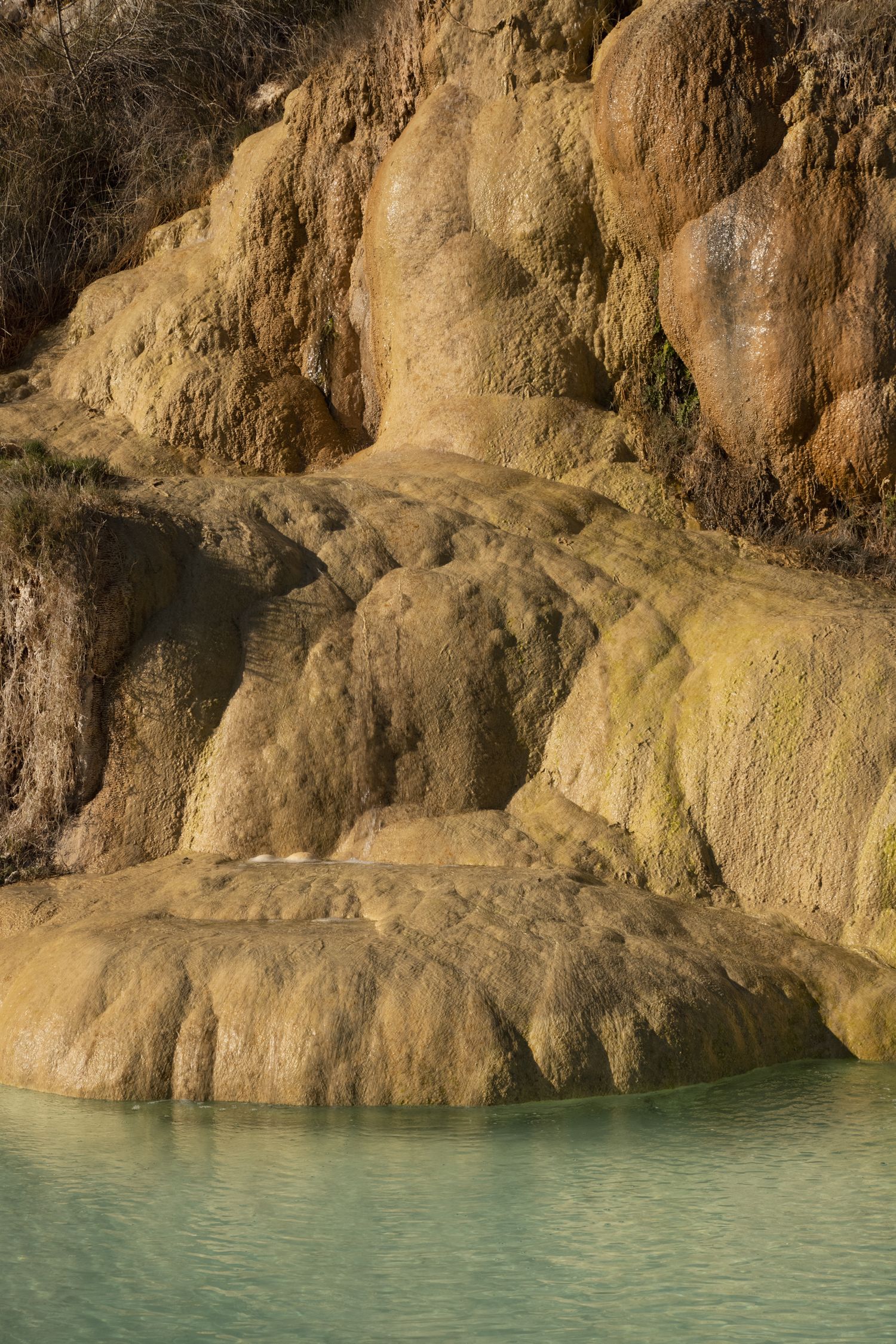 Turquoise thermal pool at base of layered travertine rock formations with mineral deposits and algae growth.