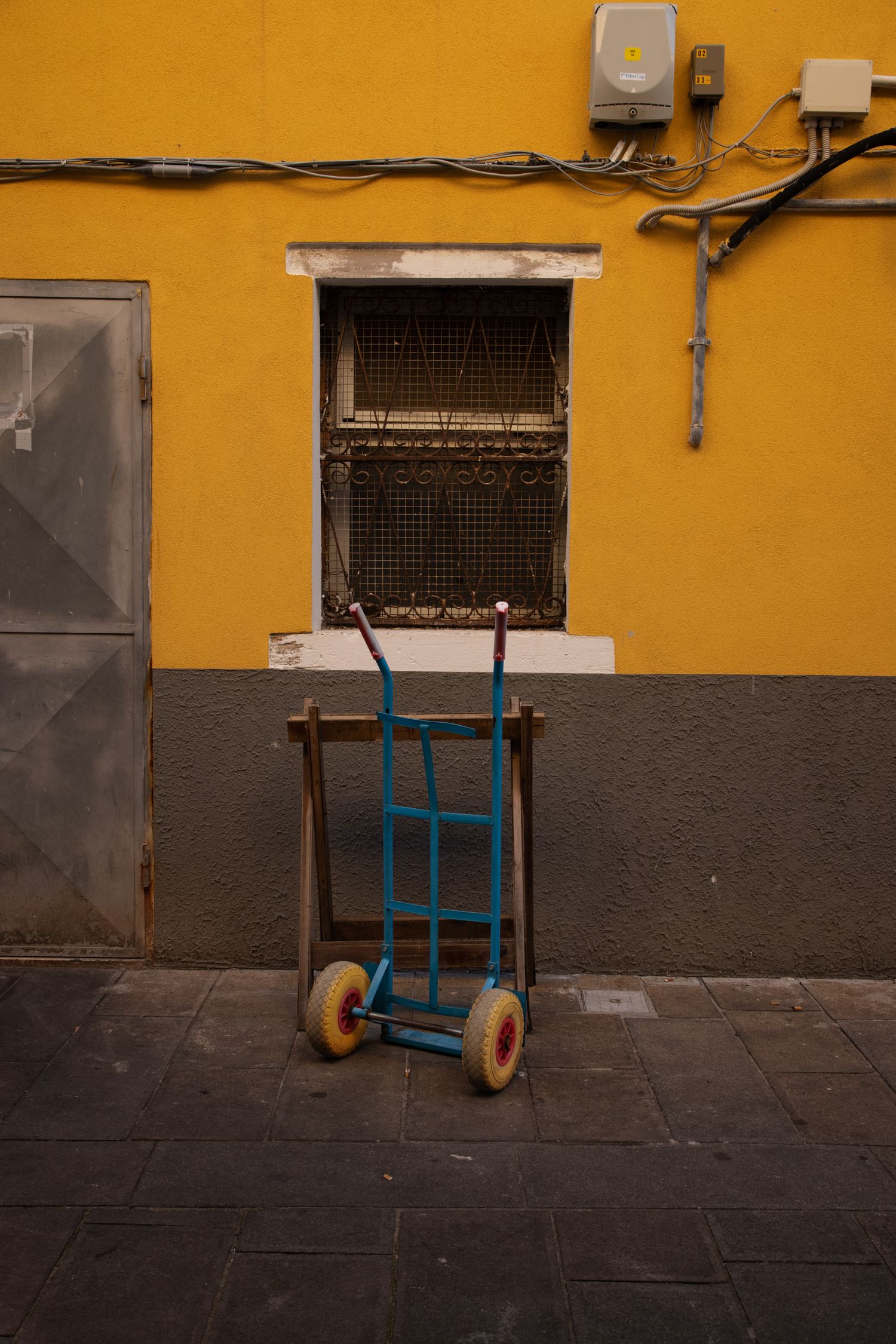 A blue hand truck rests against a yellow and gray wall beside a barred window on a cobblestone street.