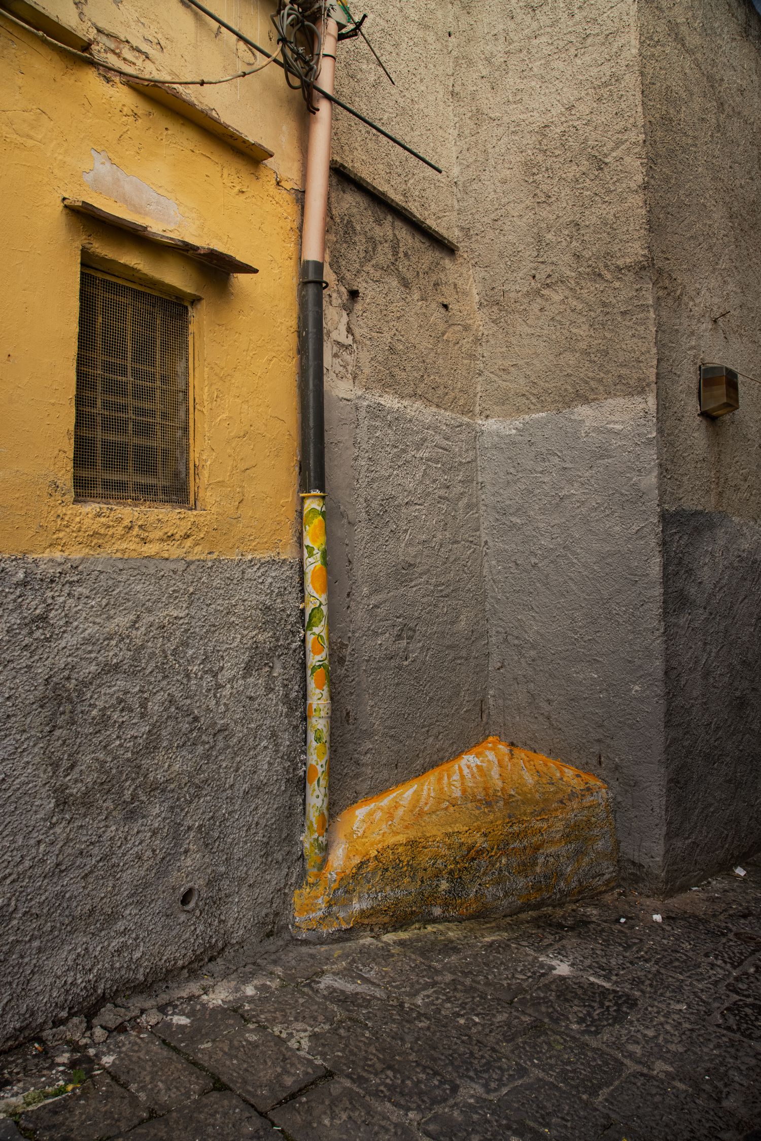 Narrow alley with yellow and gray textured walls, a drainpipe with stickers, barred window, and cobblestone ground.