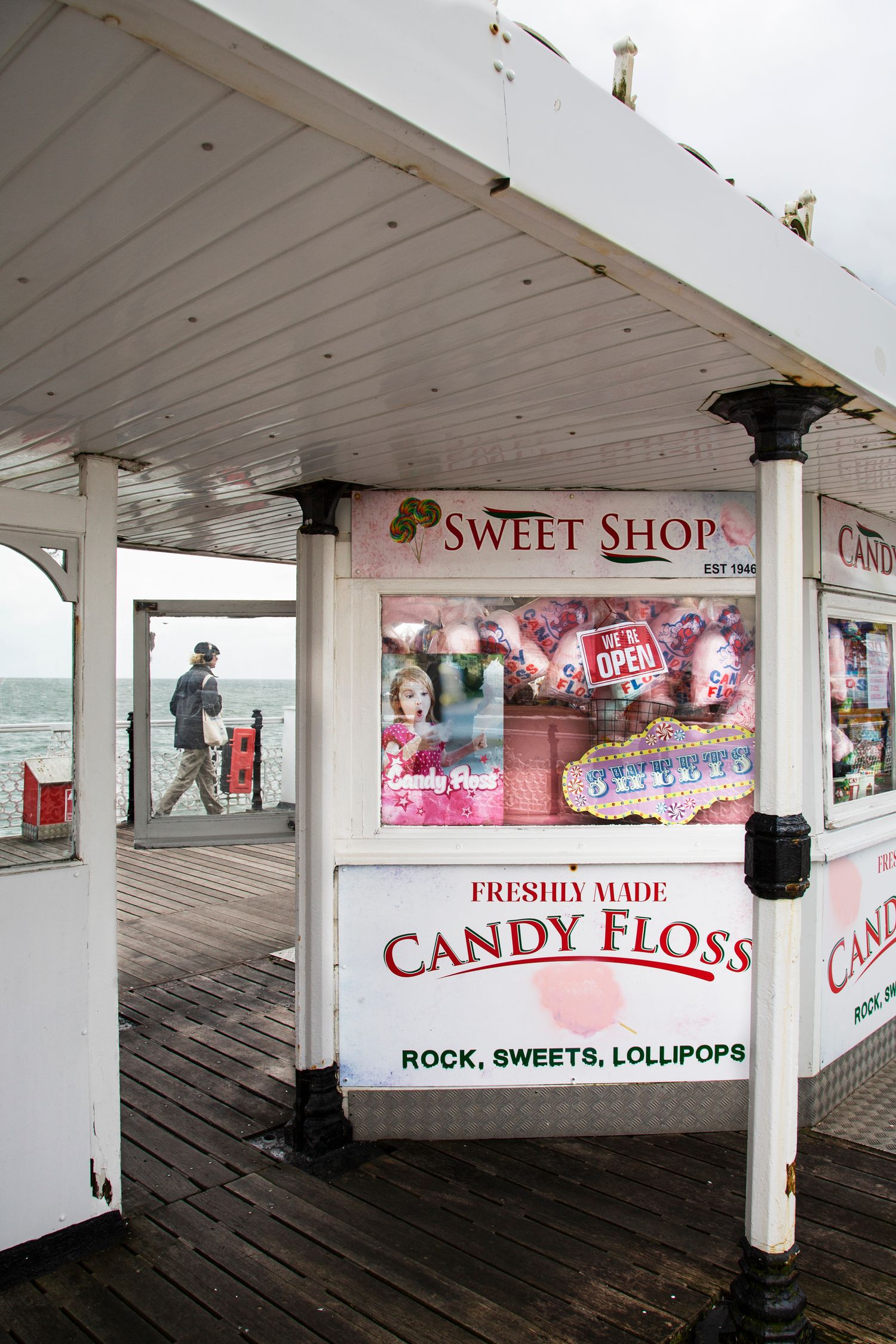 A seaside sweet shop kiosk selling candy floss and treats on a wooden pier with ocean views.
